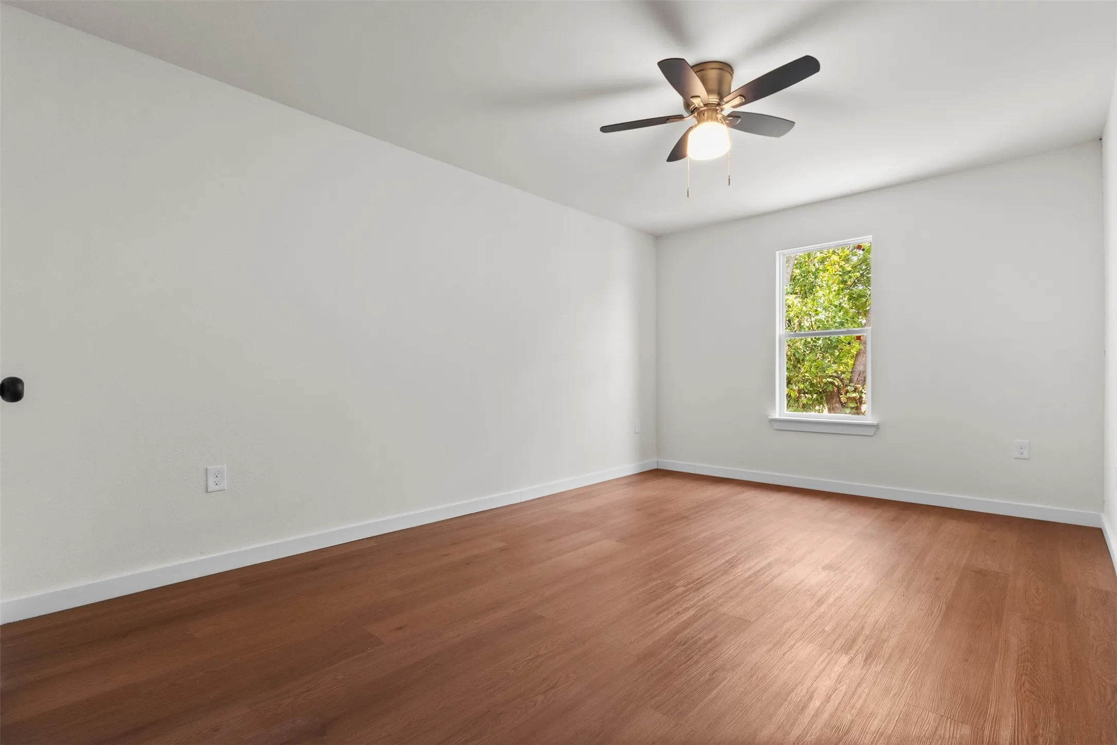 Spacious room featuring wood-finish flooring, a ceiling fan with integrated lighting, and a window providing natural illumination
