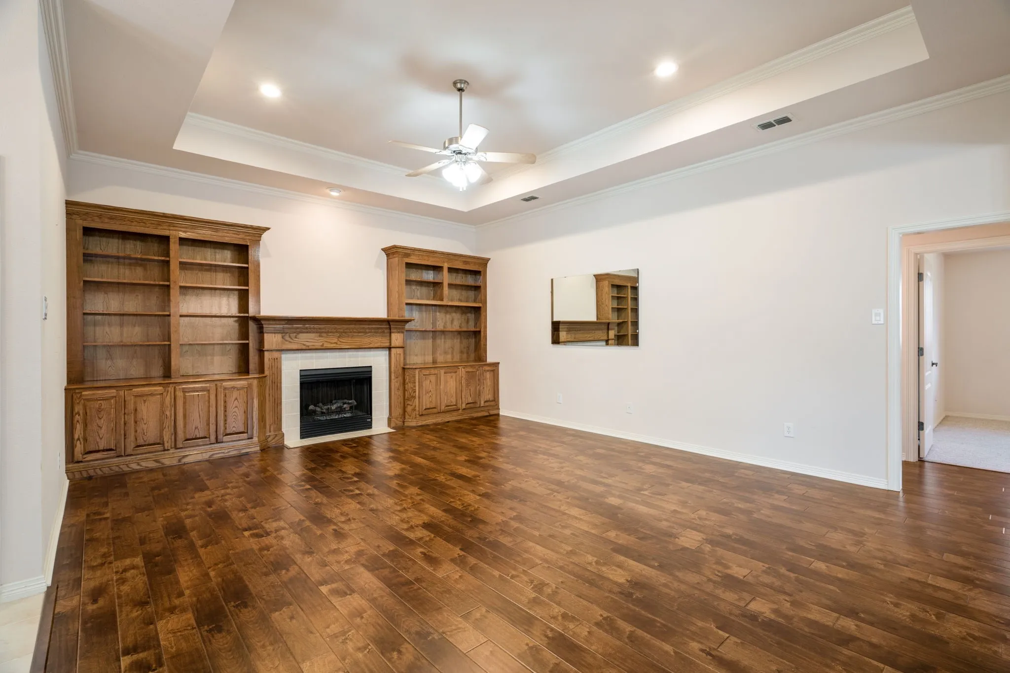 Wood-finish flooring, built-in cabinetry, fireplace with a white surround, tray ceiling, and a ceiling fan with integrated lighting