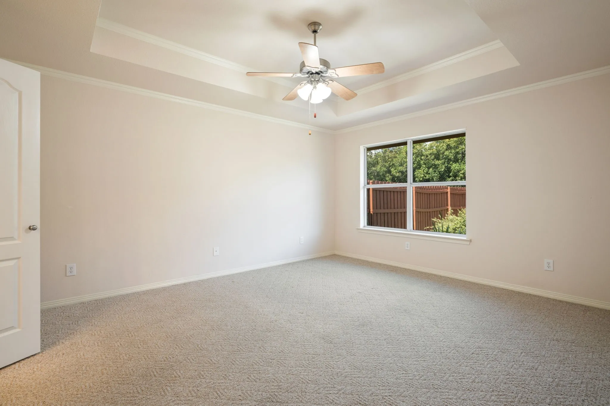 Recessed tray ceiling with crown molding, ceiling fan with integrated lighting, carpeted flooring, and a large window