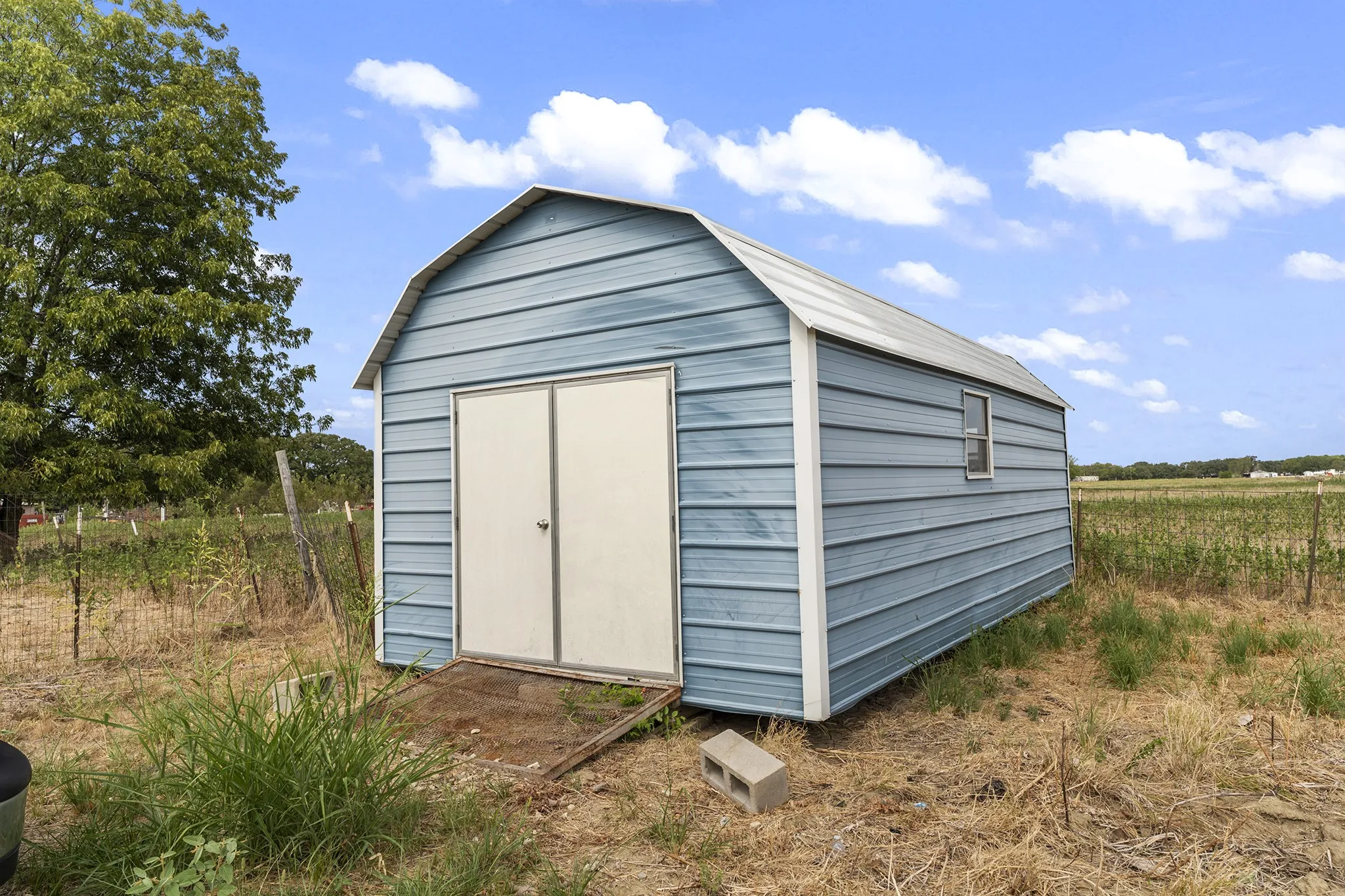 Blue metal storage shed with gambrel roof, double entry doors, and a single window