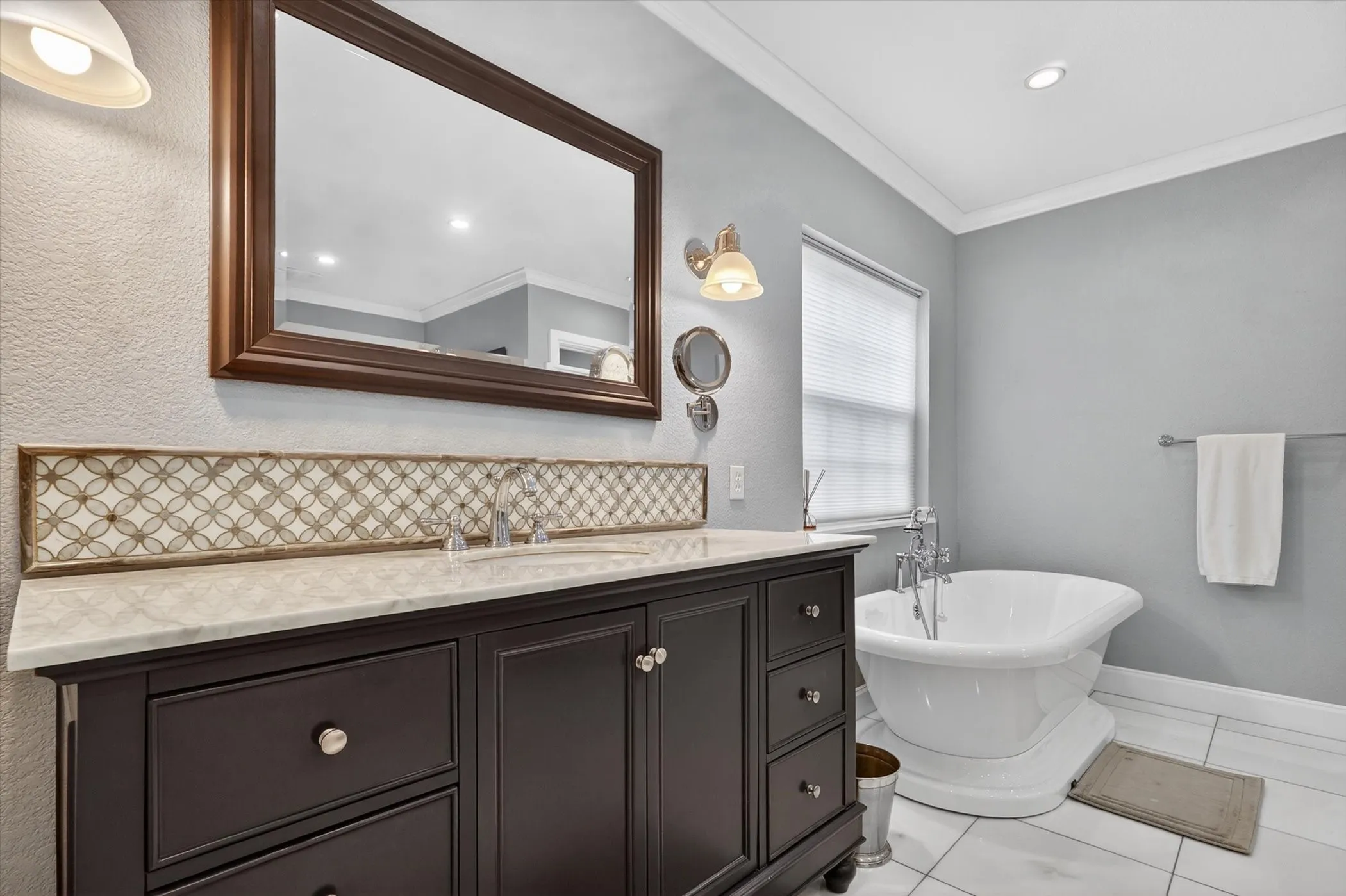 Freestanding pedestal bathtub complemented by a dark wood vanity with a light-toned countertop, a patterned tile backsplash, and a framed mirror