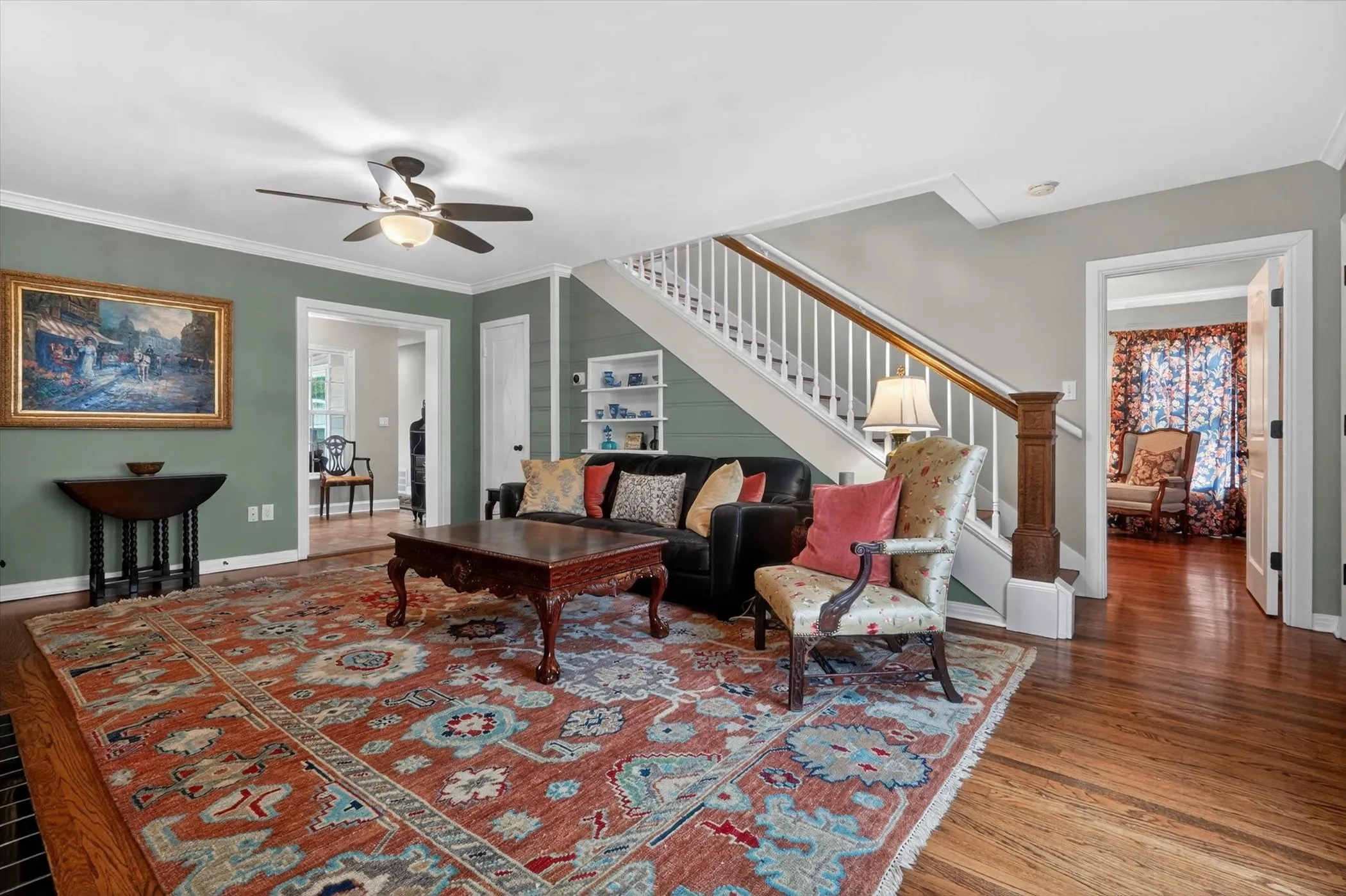 Hardwood flooring in a living space featuring a ceiling fan, crown molding, a built-in staircase, and a doorway leading to an additional room