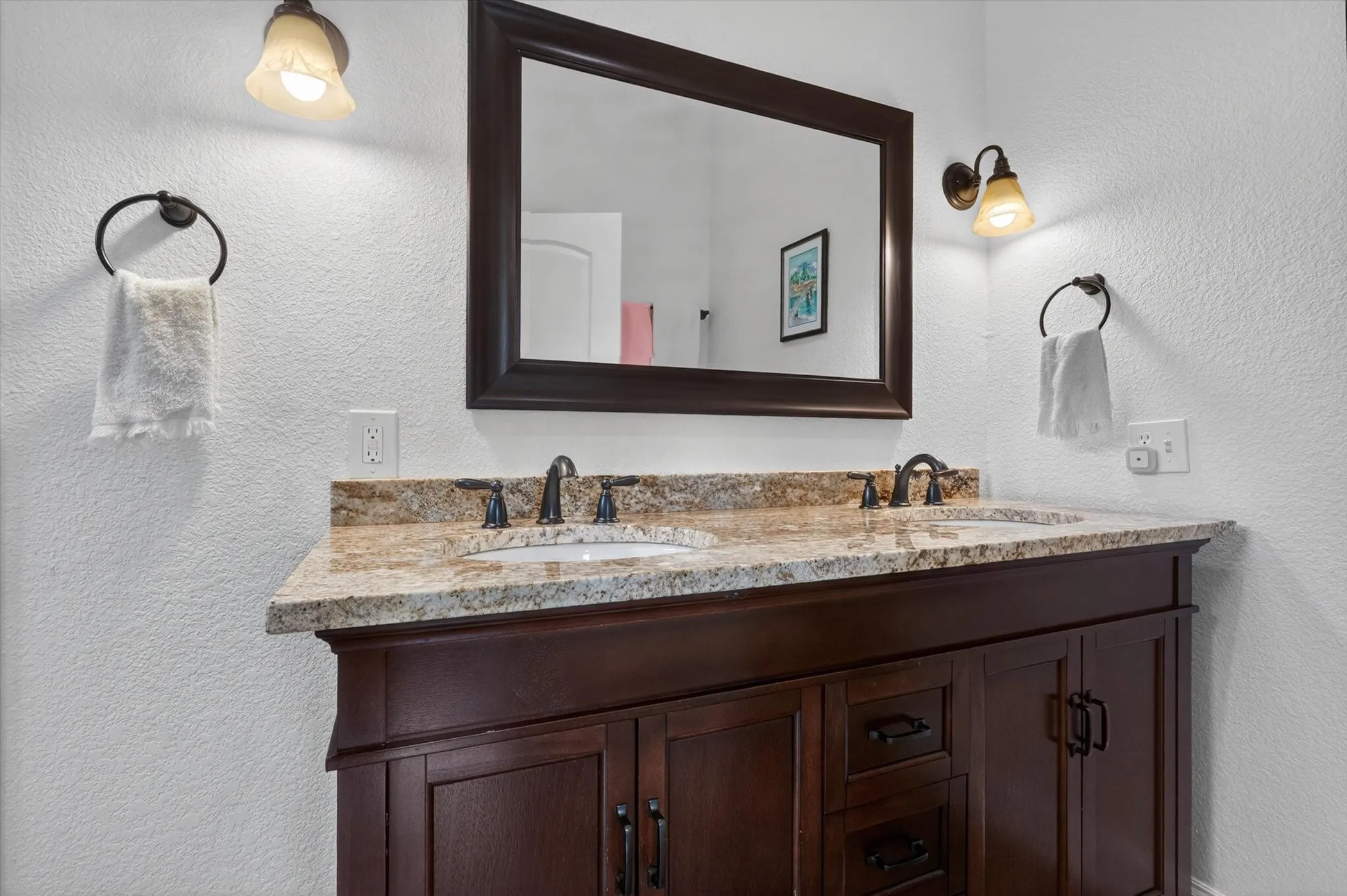 Dual sink vanity with dark wood-finish cabinetry, a patterned countertop, and bronze-finish fixtures