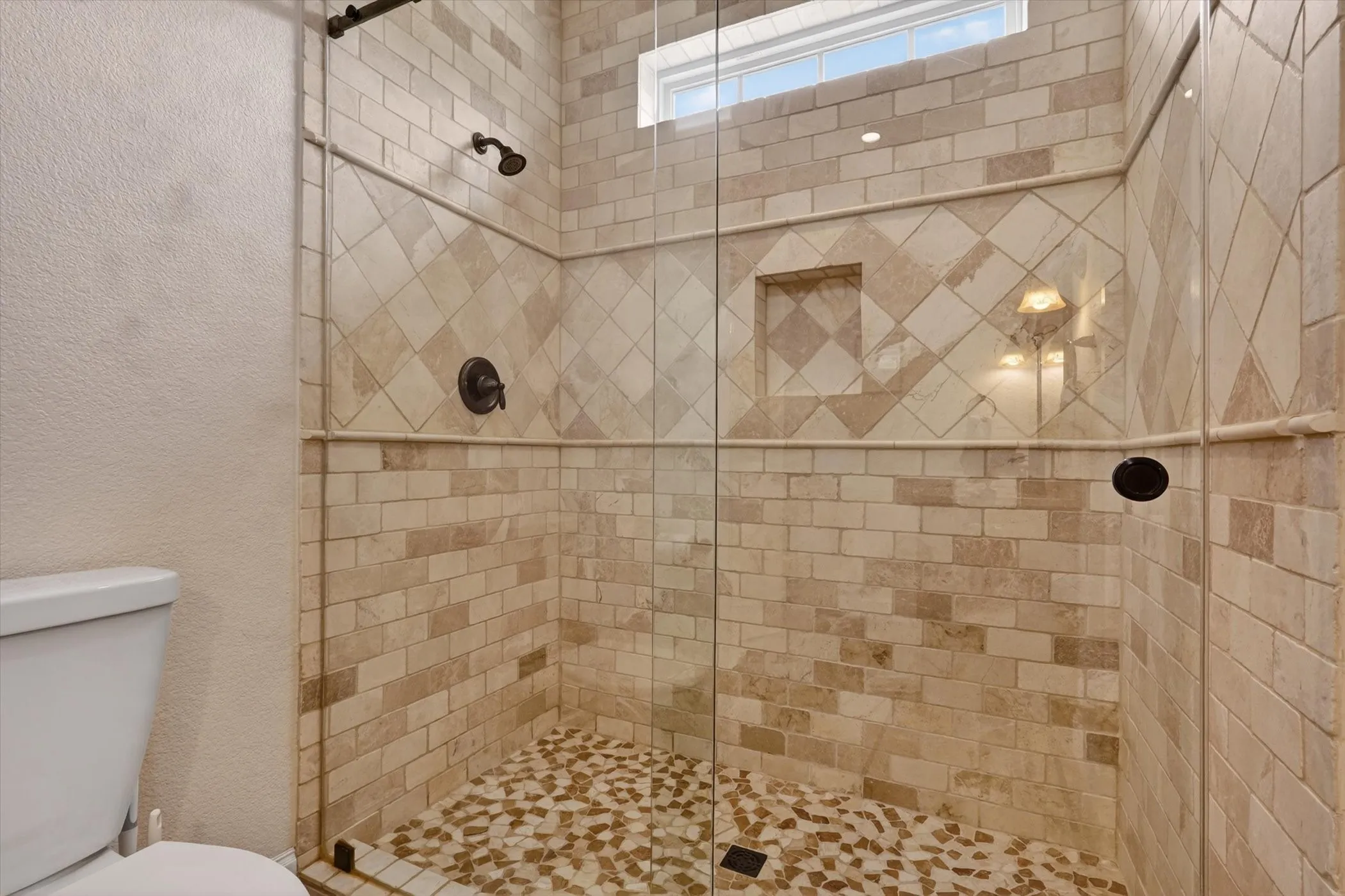 Walk-in shower featuring glass enclosure, stone-look tile walls with decorative accents, an integrated niche, dual shower heads, and a pebble-finish floor