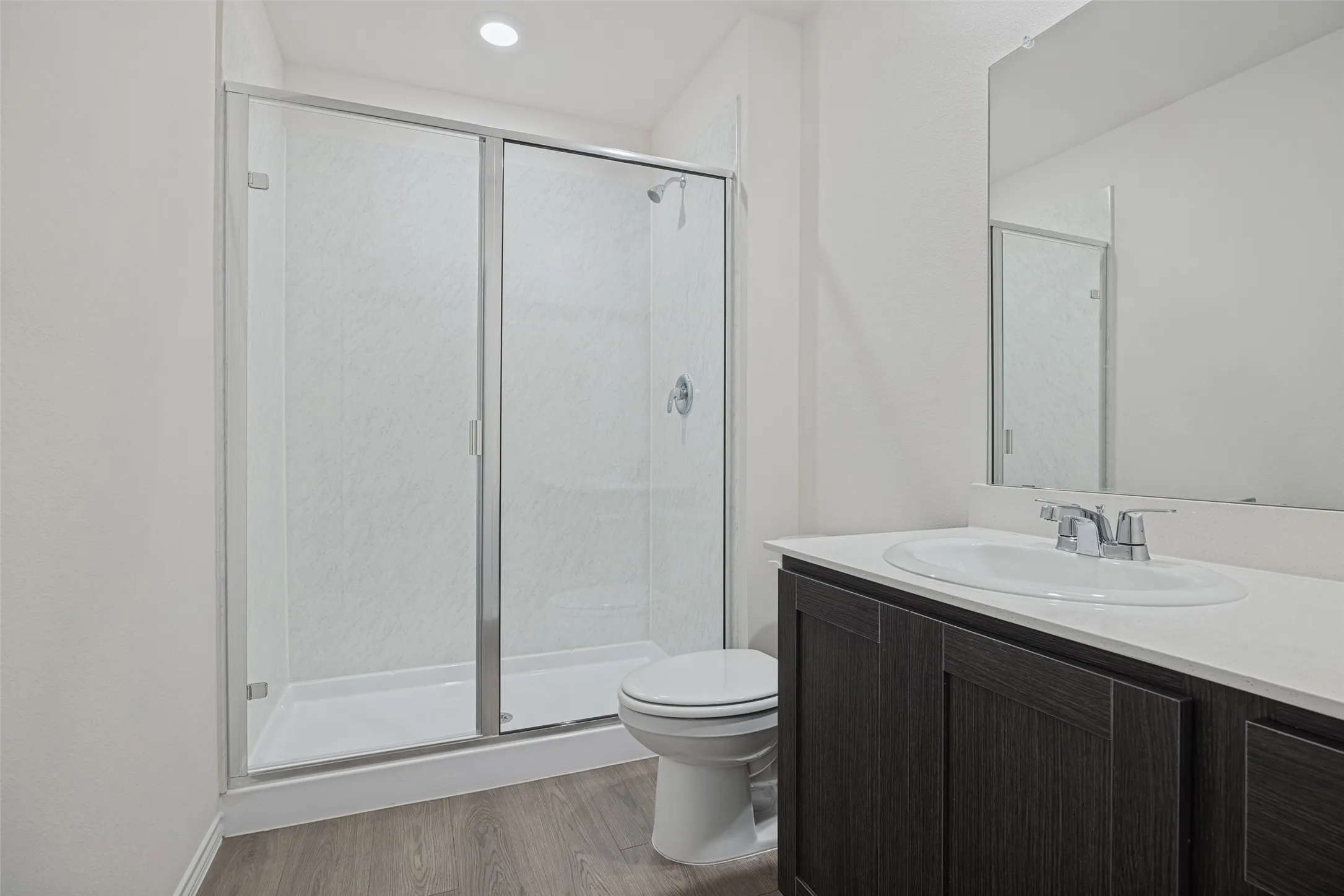 Modern bathroom featuring a frameless glass-enclosed shower with subway tile surround, dual vanity with dark wood cabinetry and white countertop, large mirror with integrated lighting, ceramic toilet, and wood-finish flooring throughout