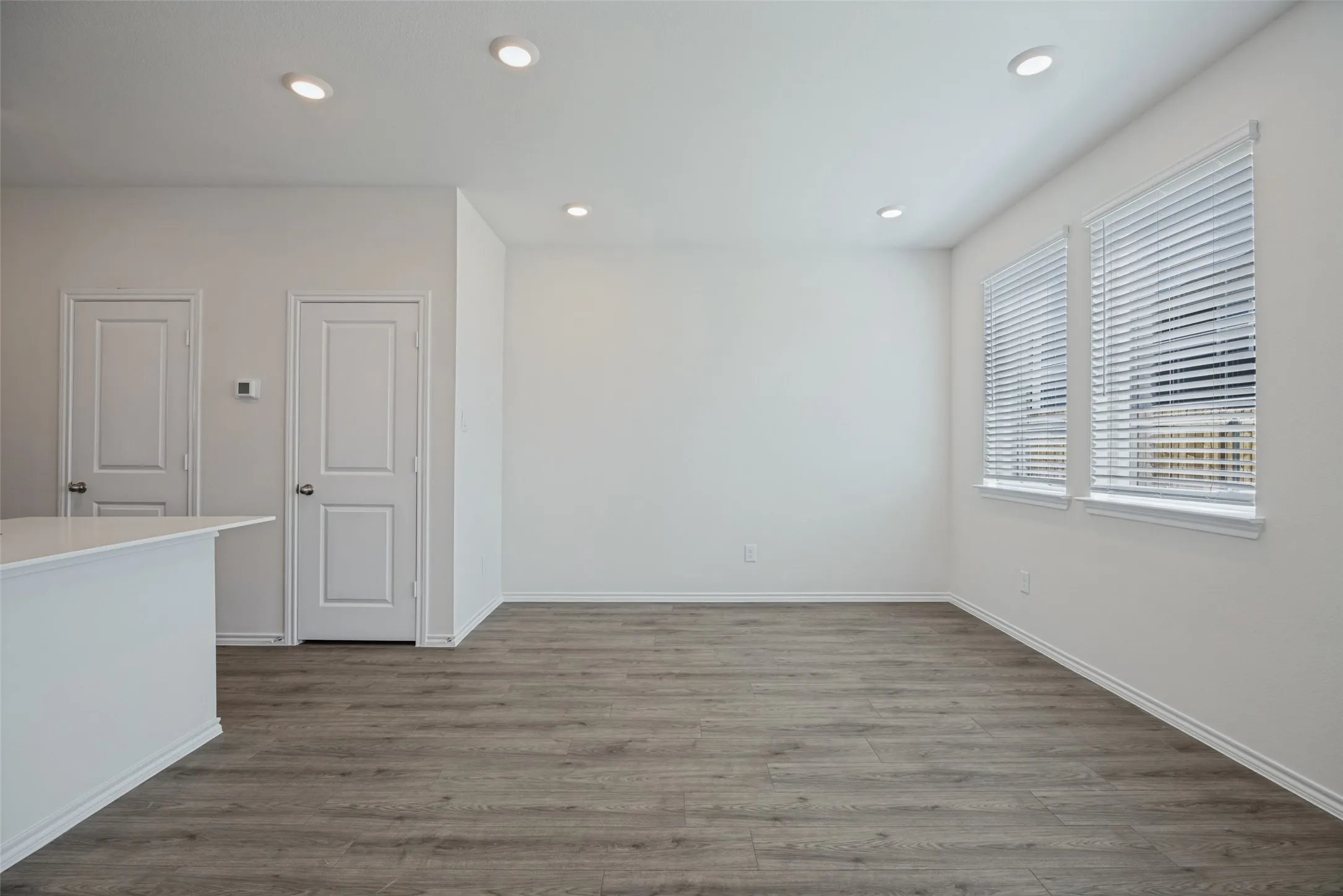 Recessed ceiling lighting, wood-finish flooring, two panel doors, white window blinds, and a kitchen island with a white countertop