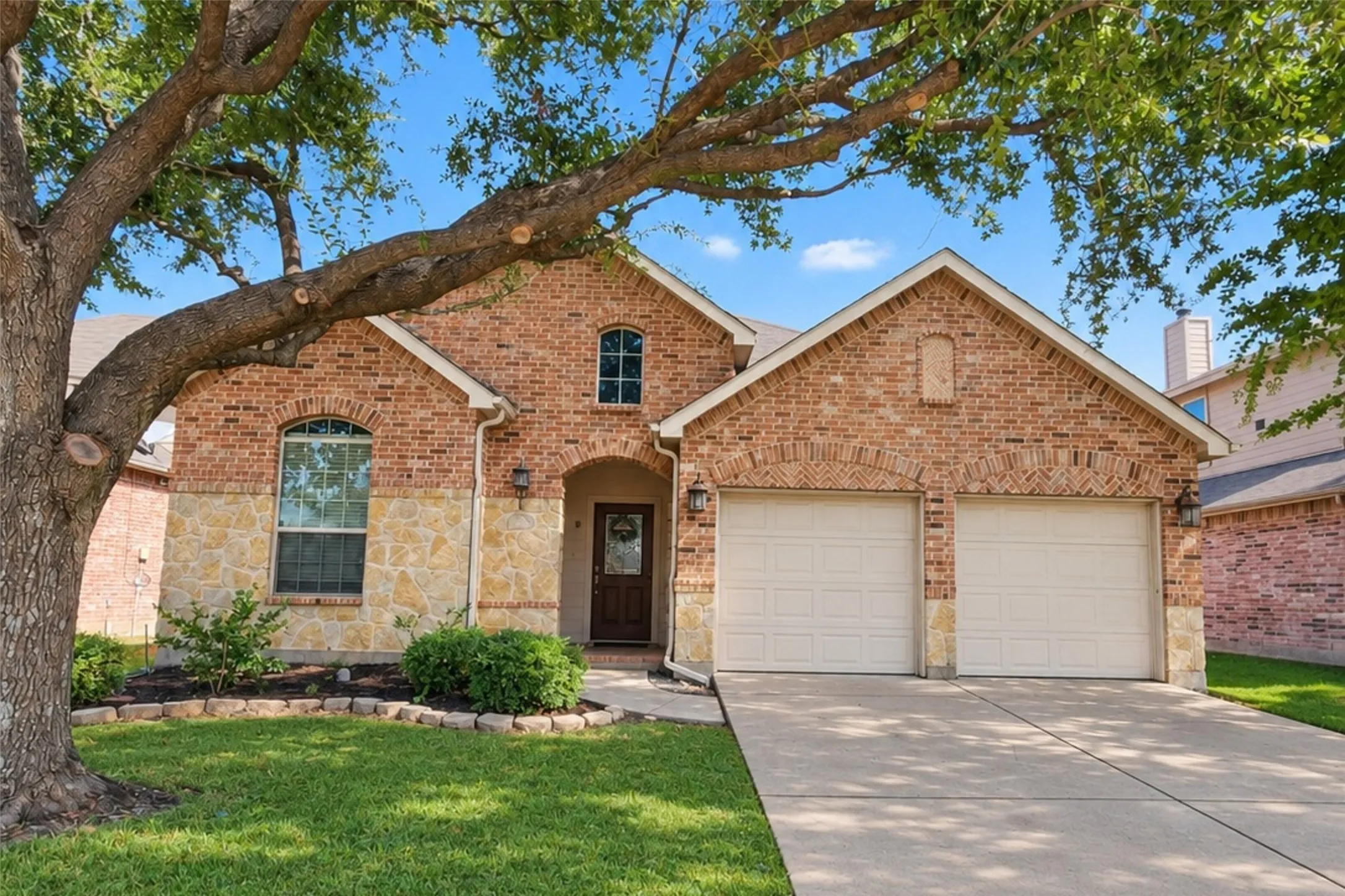 Brick and stone veneer exterior with arched window details, a two-car garage, and mature front yard tree