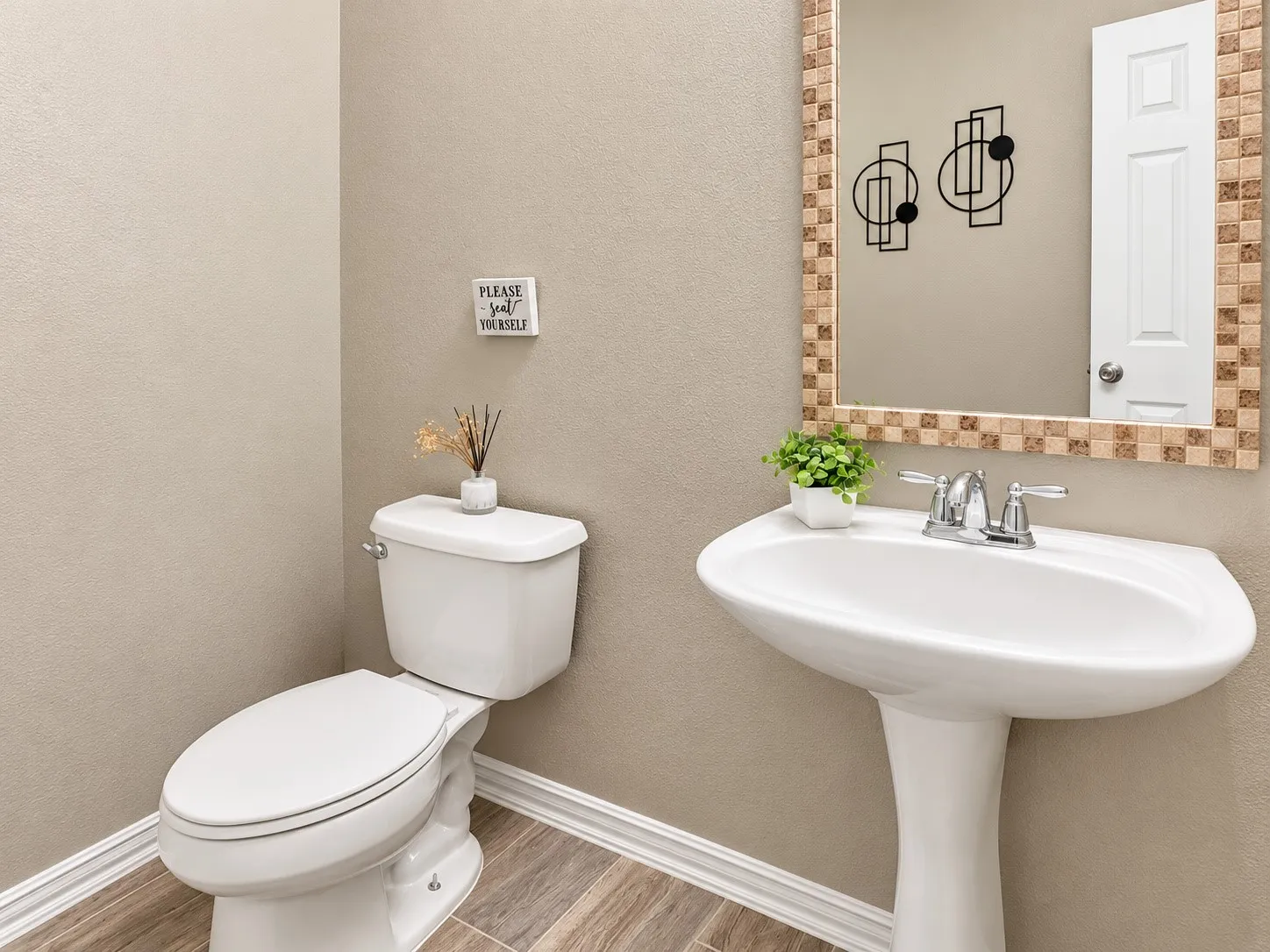 Pedestal sink with chrome faucet, toilet, wood-finish tile flooring, and a decorative mirror with a mosaic tile frame
