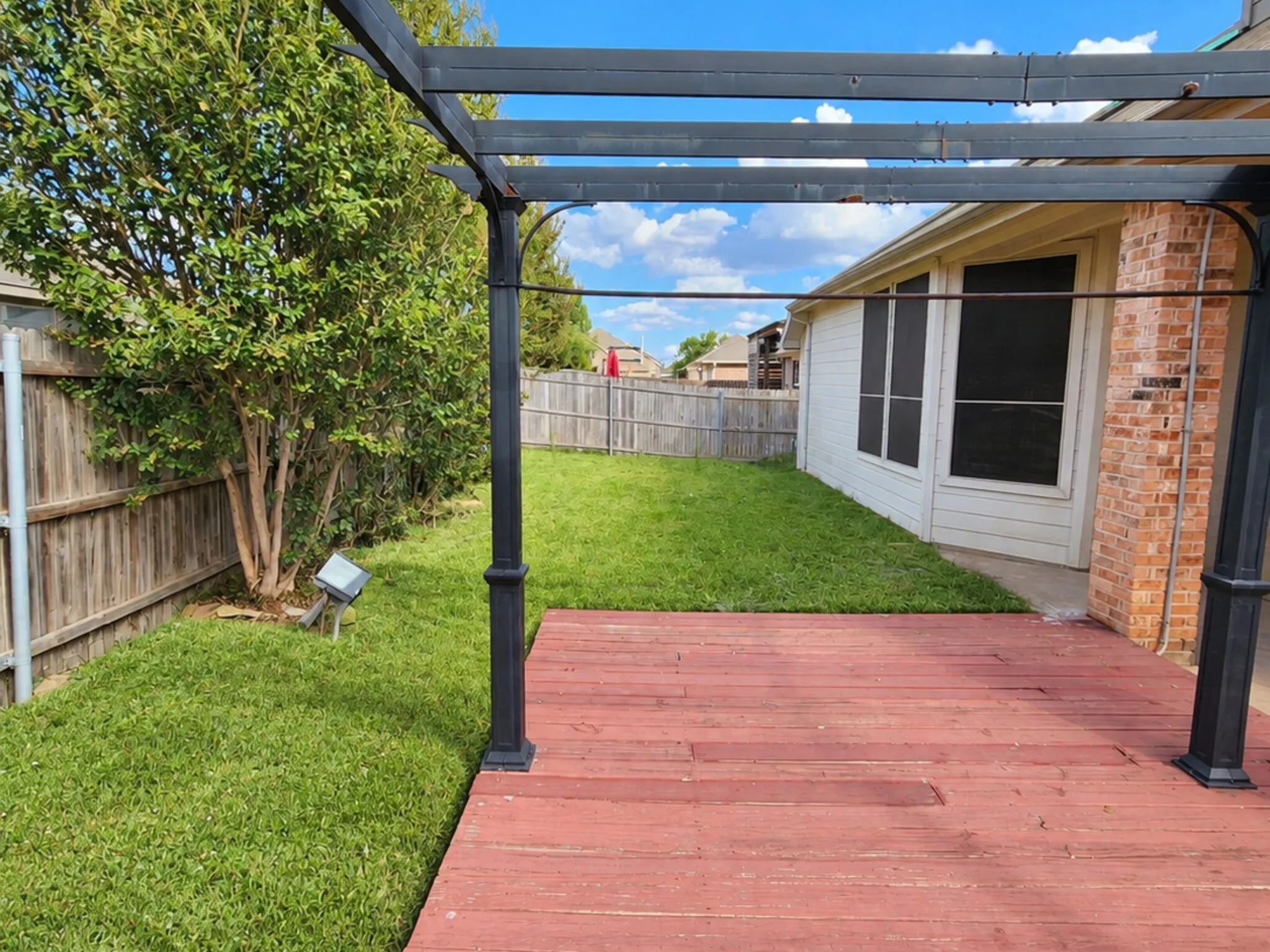Backyard featuring a red-stained wood deck with a black metal pergola, a manicured lawn, and a wooden privacy fence