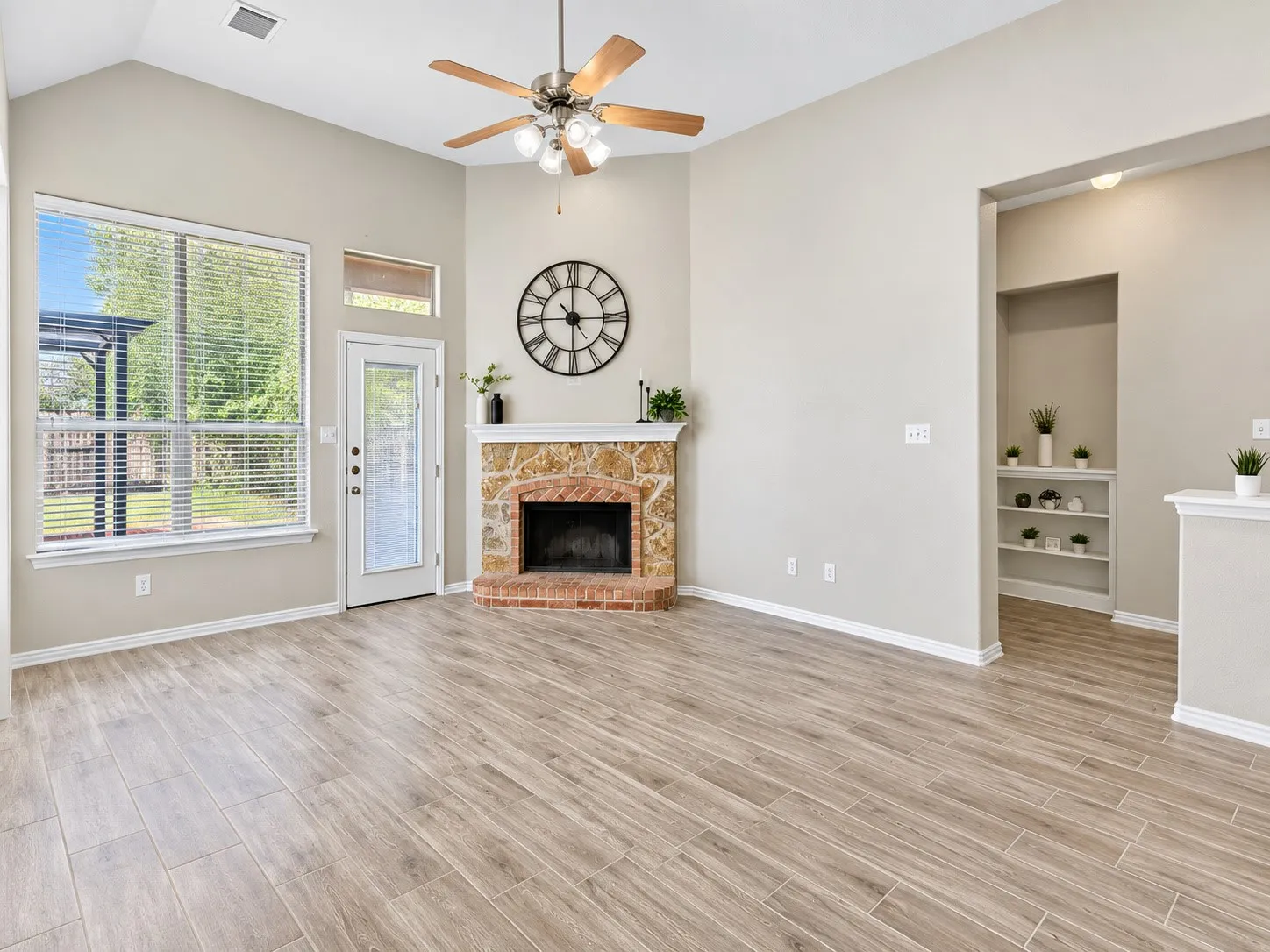 Vaulted ceiling with a ceiling fan, wood-finish flooring, corner fireplace with stone and brick surround, large window, and built-in shelving