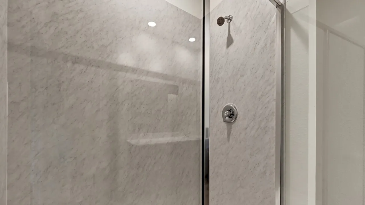 Glass-enclosed shower with a polished chrome shower head and control, featuring light-toned, textured wall panels and integrated ceiling lighting