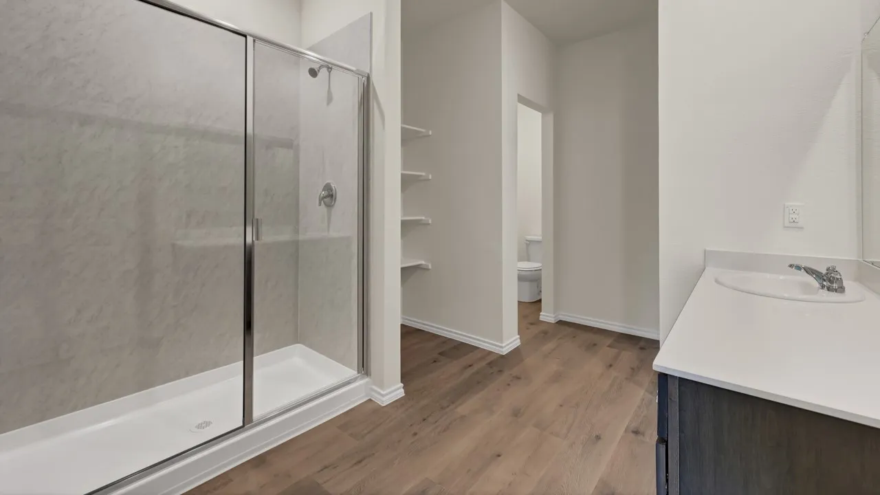 Walk-in shower with sliding glass enclosure, white vanity with integrated sink and chrome faucet, wood-finish flooring, and built-in shelving