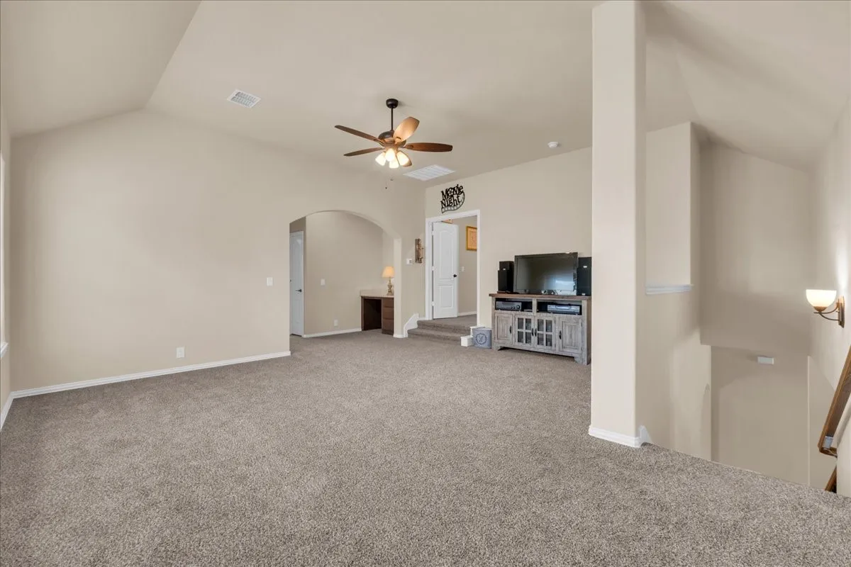 Spacious carpeted room featuring a vaulted ceiling with a ceiling fan, an arched doorway, and built-in wall sconces