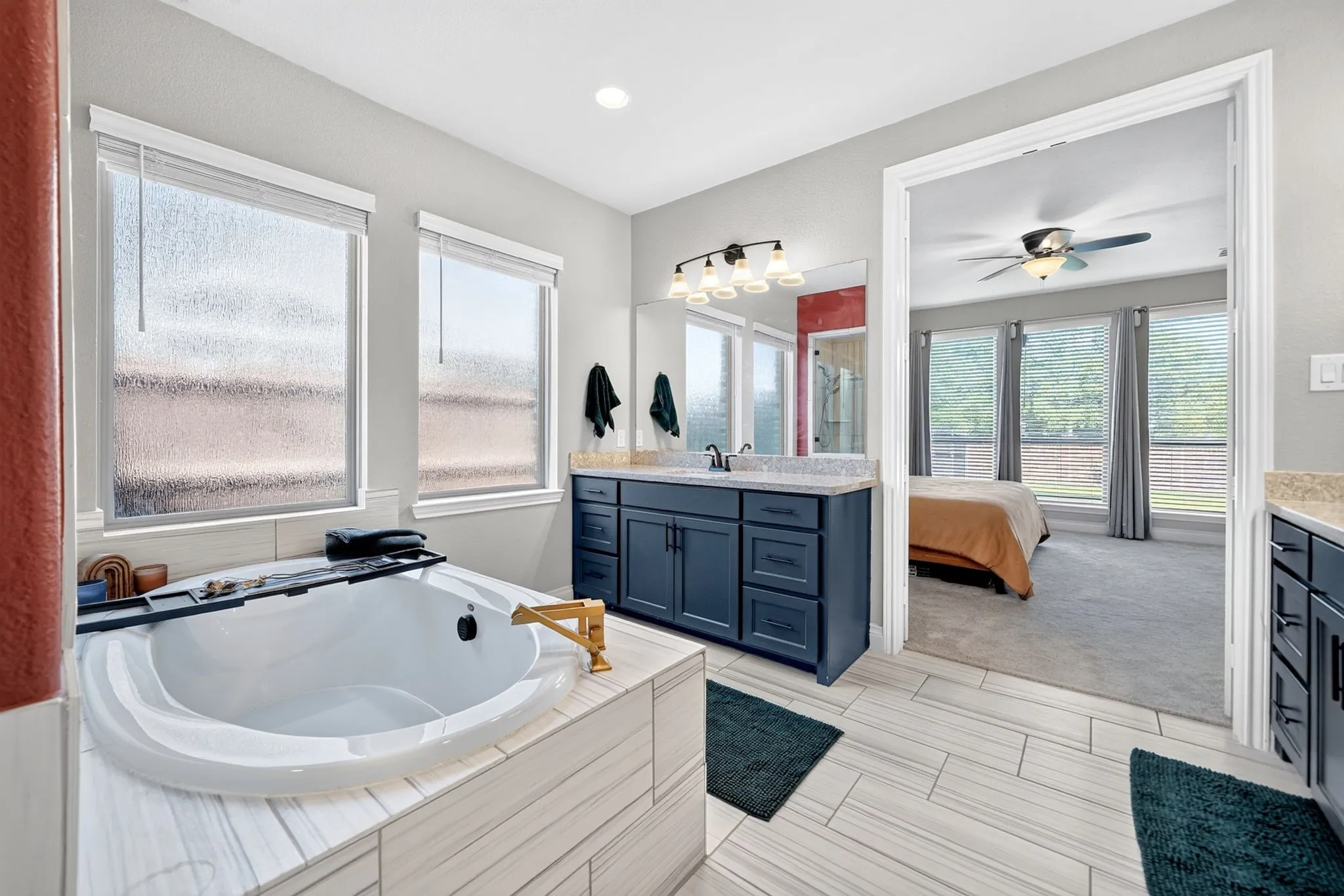 Soaking tub with a gold-finish faucet, dual-sink vanity with a light-colored countertop, large framed mirror, and light-toned tile​​‌​​​​‌​​‌‌​‌‌​​​‌‌​‌​‌​‌​​​‌​​ flooring