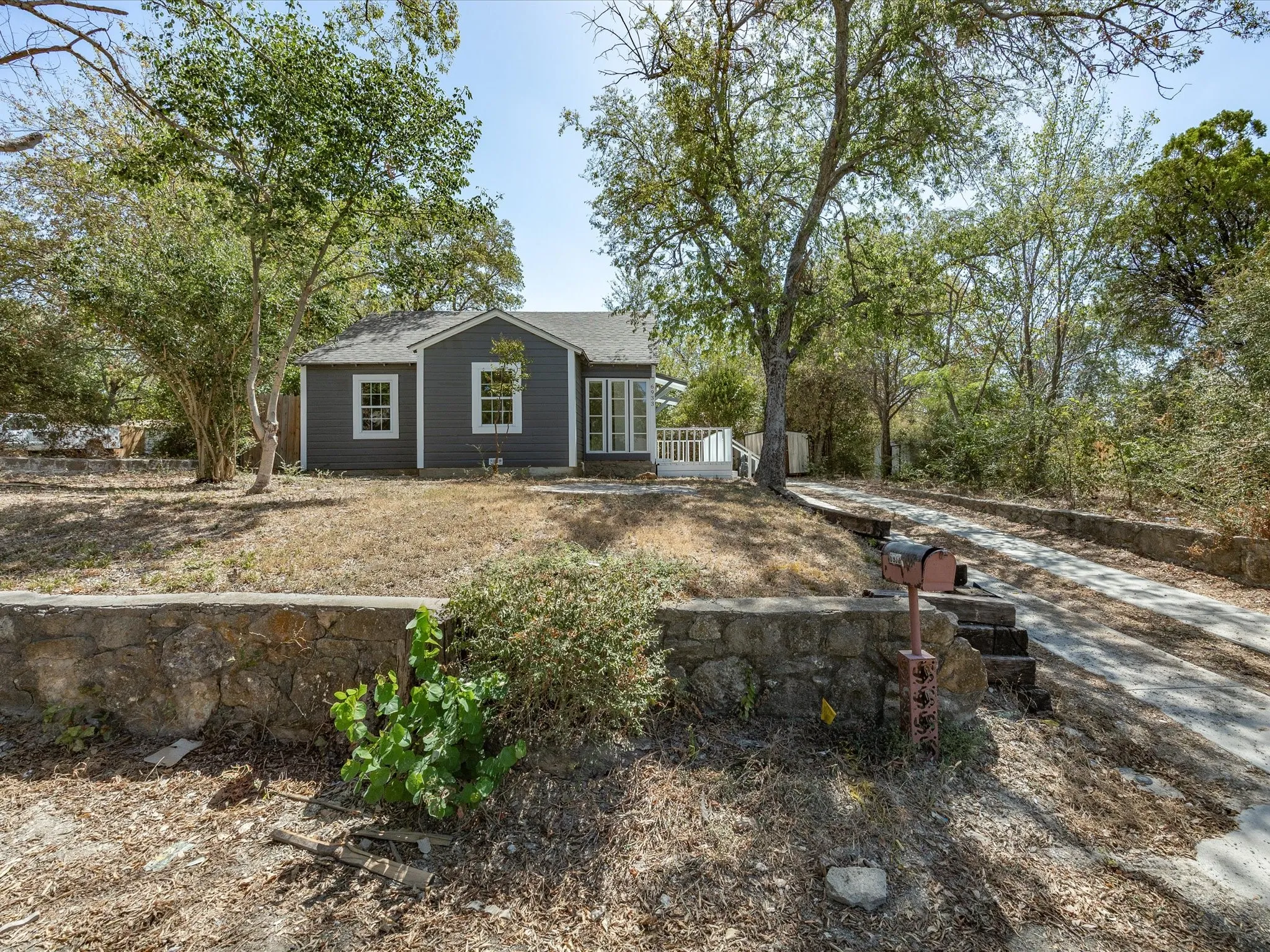 Dark gray siding exterior with white window trim, multiple mature trees, a paved driveway, and a stone retaining wall