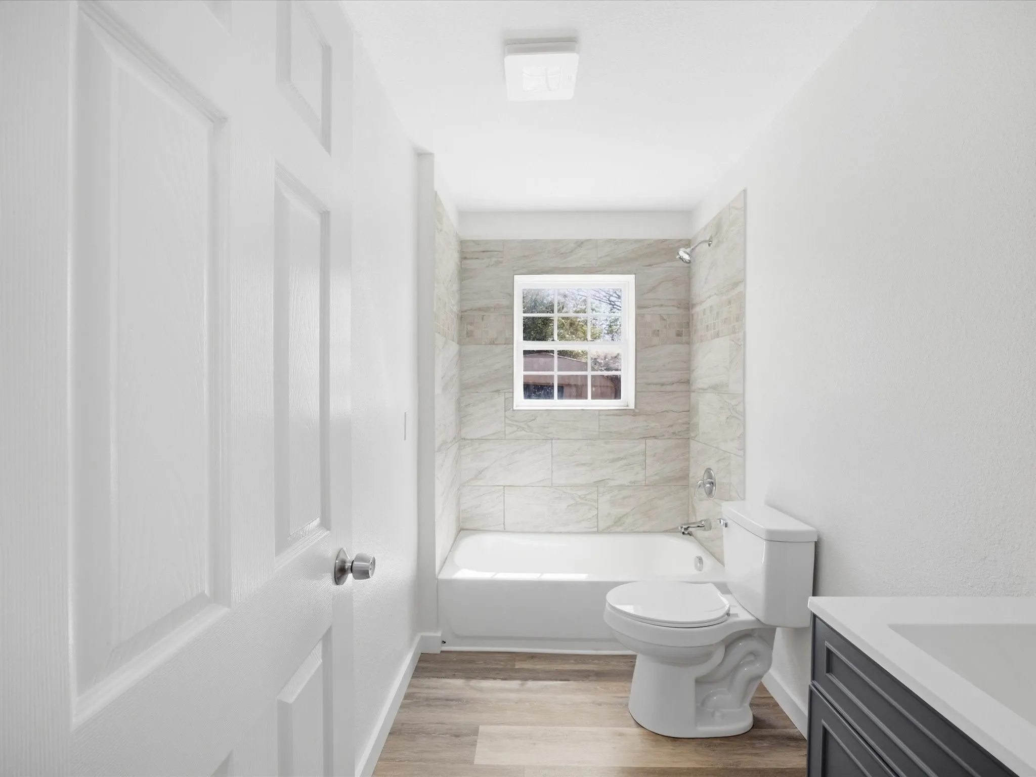 Bathroom featuring a windowed shower-tub combination with full-height tile surround, wood-finish flooring, a white toilet, and a vanity with a white countertop