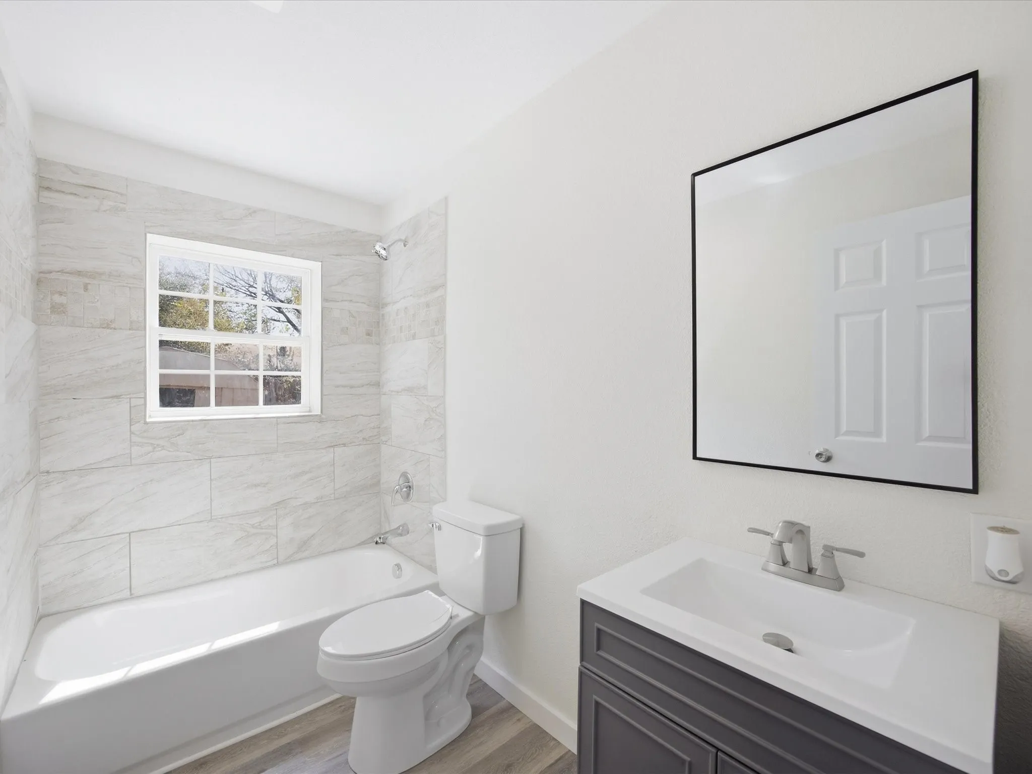 Bathroom featuring a vanity with a rectangular sink, a framed mirror, a bathtub with tile surround, and wood-finish flooring
