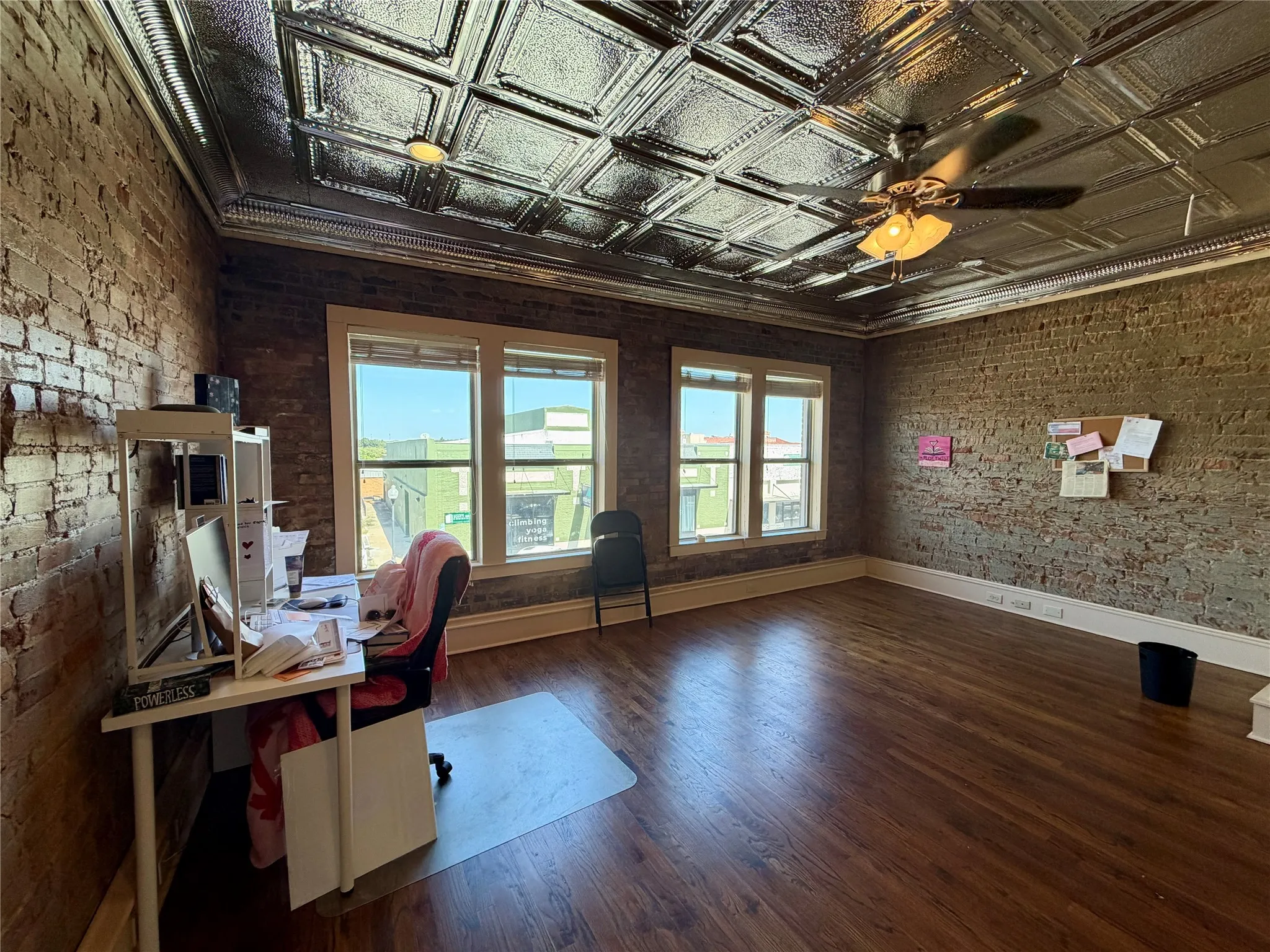 Exposed brick walls, wood-finish flooring, pressed metal ceiling, multiple windows, and a ceiling fan with integrated lighting