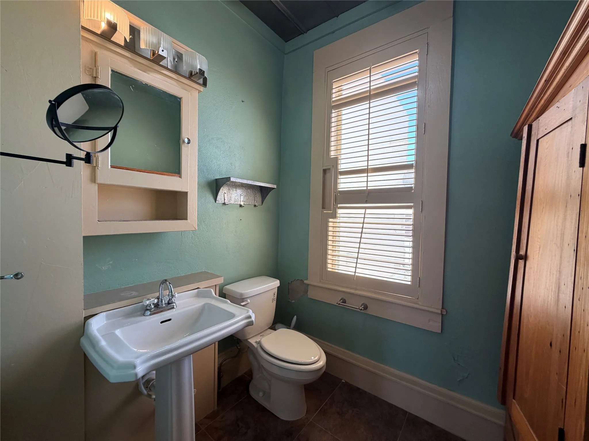 Pedestal sink with chrome faucet, wall-mounted medicine cabinet with integrated lighting, wood-finish flooring, and a large window with blinds