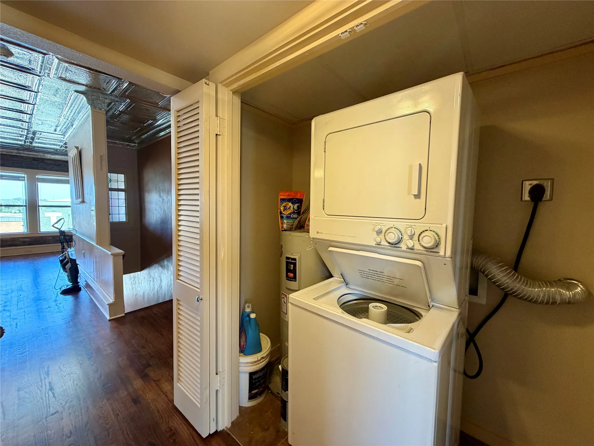 Stackable white washer and dryer unit, wood-finish flooring, white louvered bifold doors, window-lined wall, decorative ceiling panels