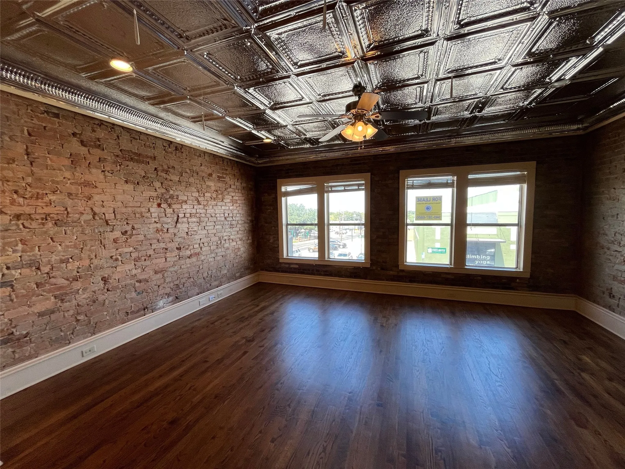 Exposed brick walls, wood-finish flooring, two large windows, tin tile ceiling, and a ceiling fan with integrated lighting