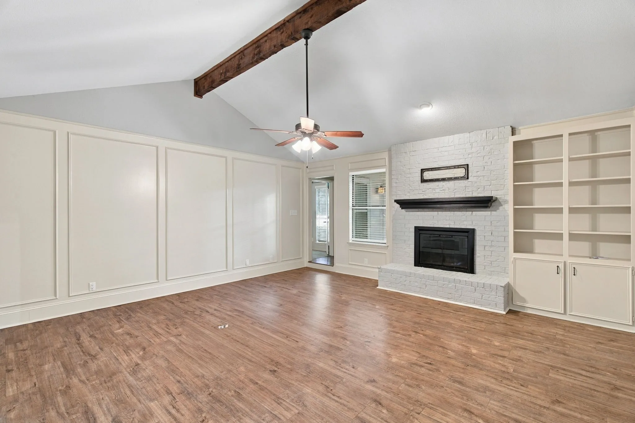 Vaulted ceiling featuring a wood beam, wood-finish flooring, white brick fireplace, built-in shelving, and decorative wall paneling
