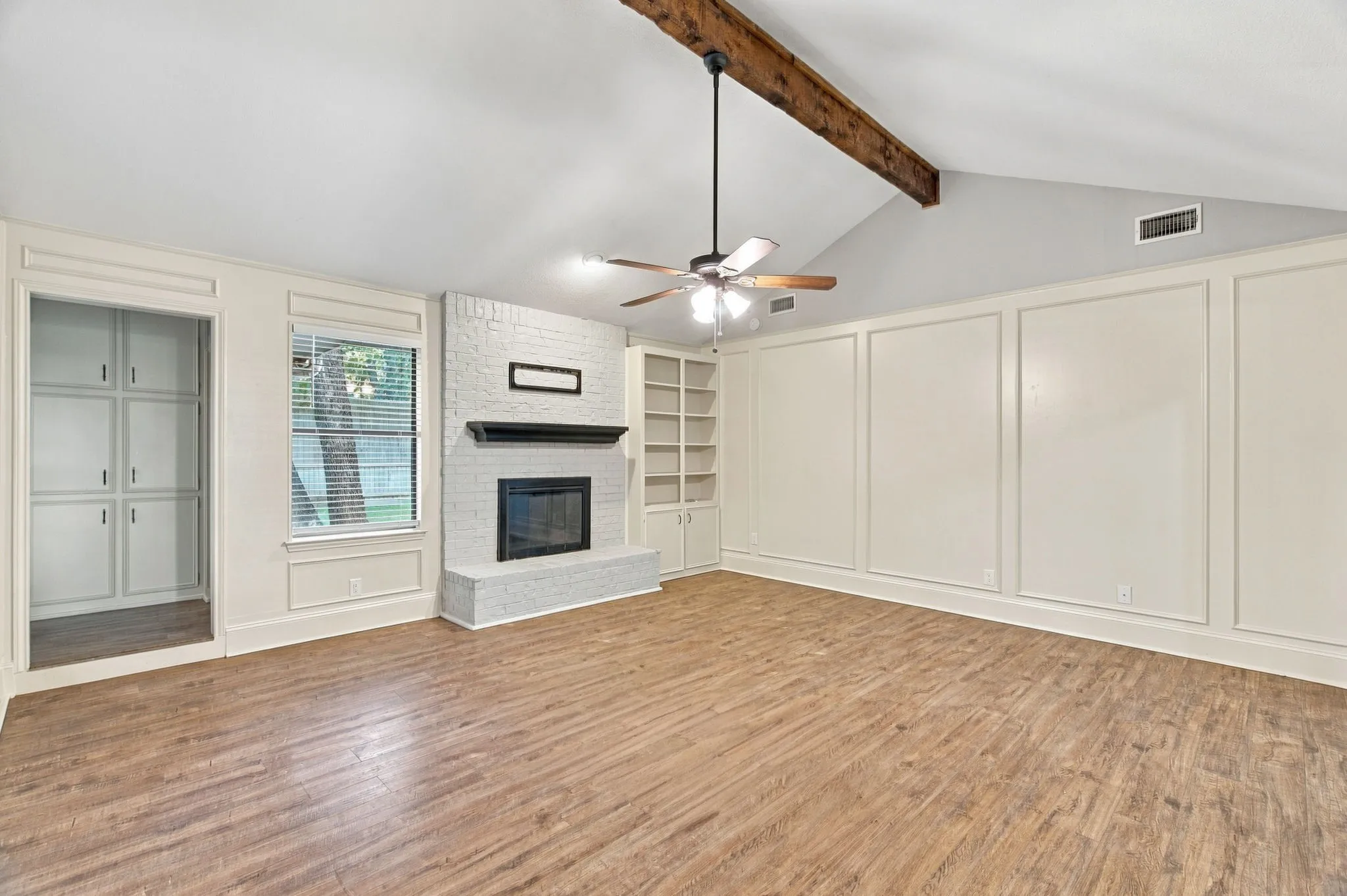 Vaulted ceiling with an exposed wood beam, wood-finish flooring, brick-surround fireplace with a dark mantel, built-in shelving, and built-in cabinetry