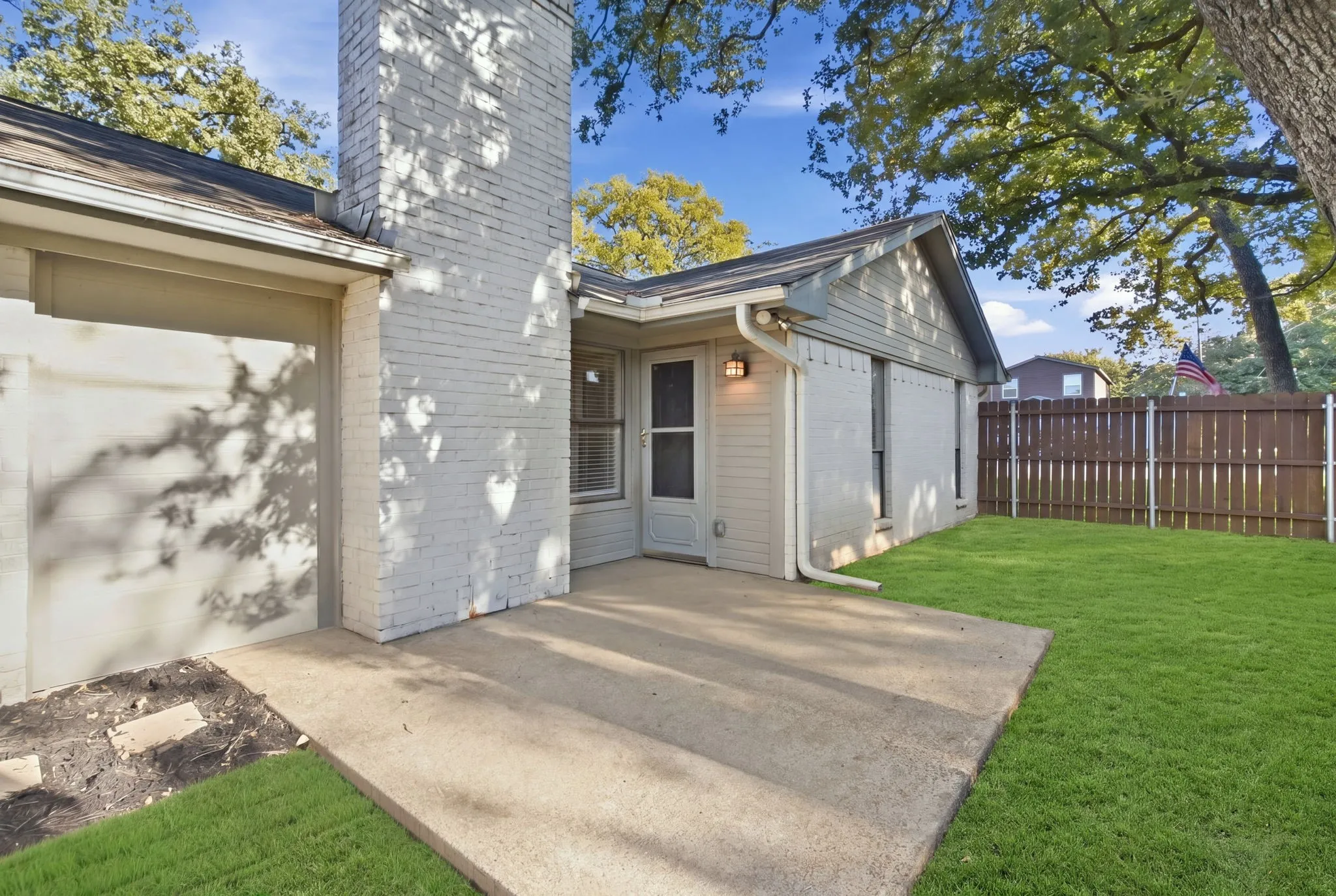 Expansive concrete patio, white brick chimney, wood fence, and established trees