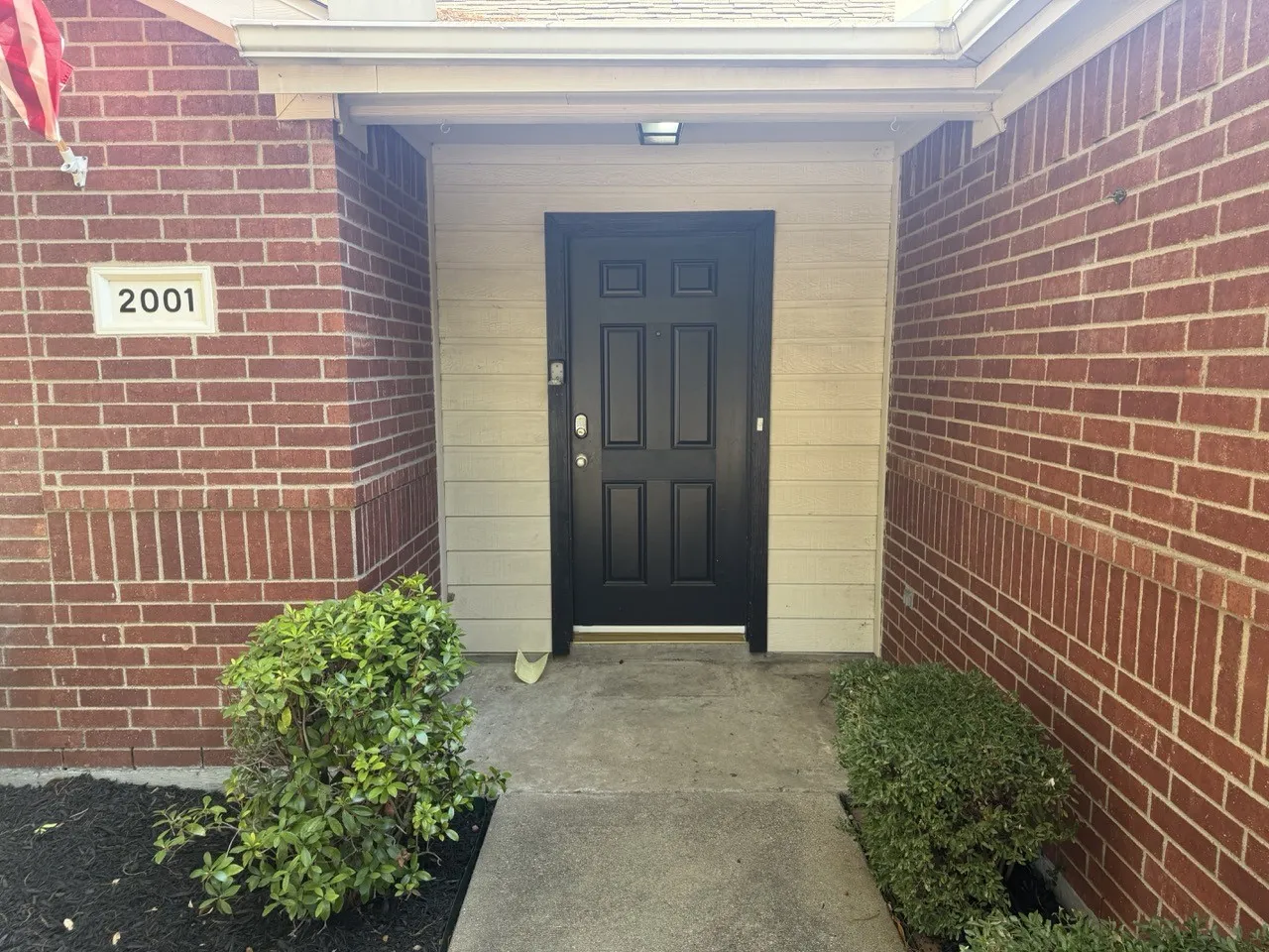 Brick exterior with a recessed entryway, featuring a panel-style black door, light-colored siding, and concrete walkway
