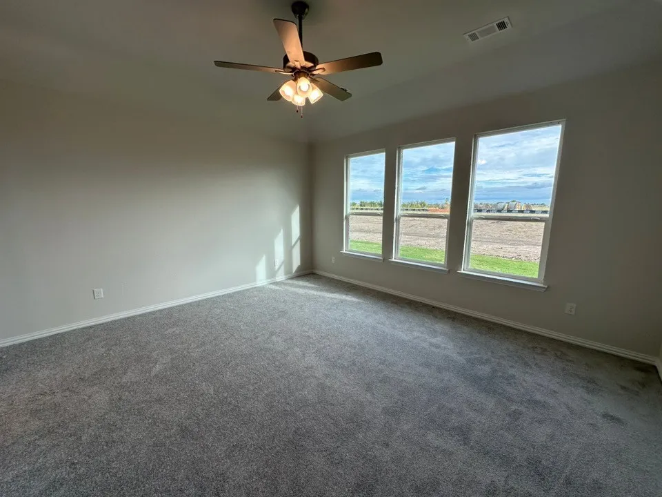 Plush gray carpeting, three large windows, neutral wall tones, and a ceiling fan with integrated lighting