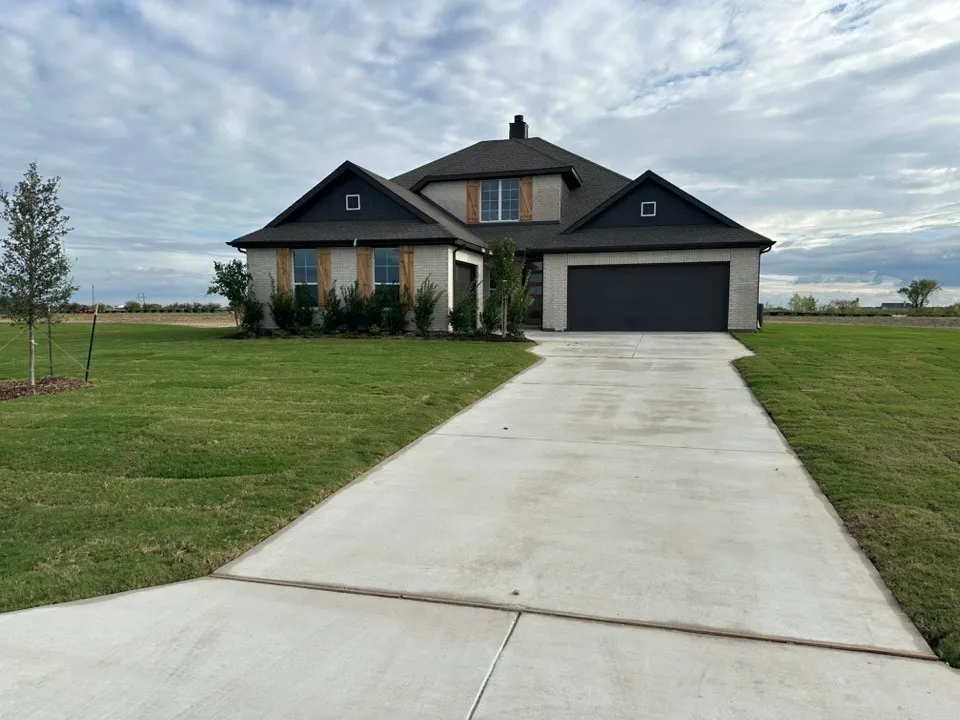 Light-colored brick exterior with dark trim accents, natural wood-finish window shutters, an attached two-car garage with a dark garage door, a prominent concrete driveway, and a manicured lawn