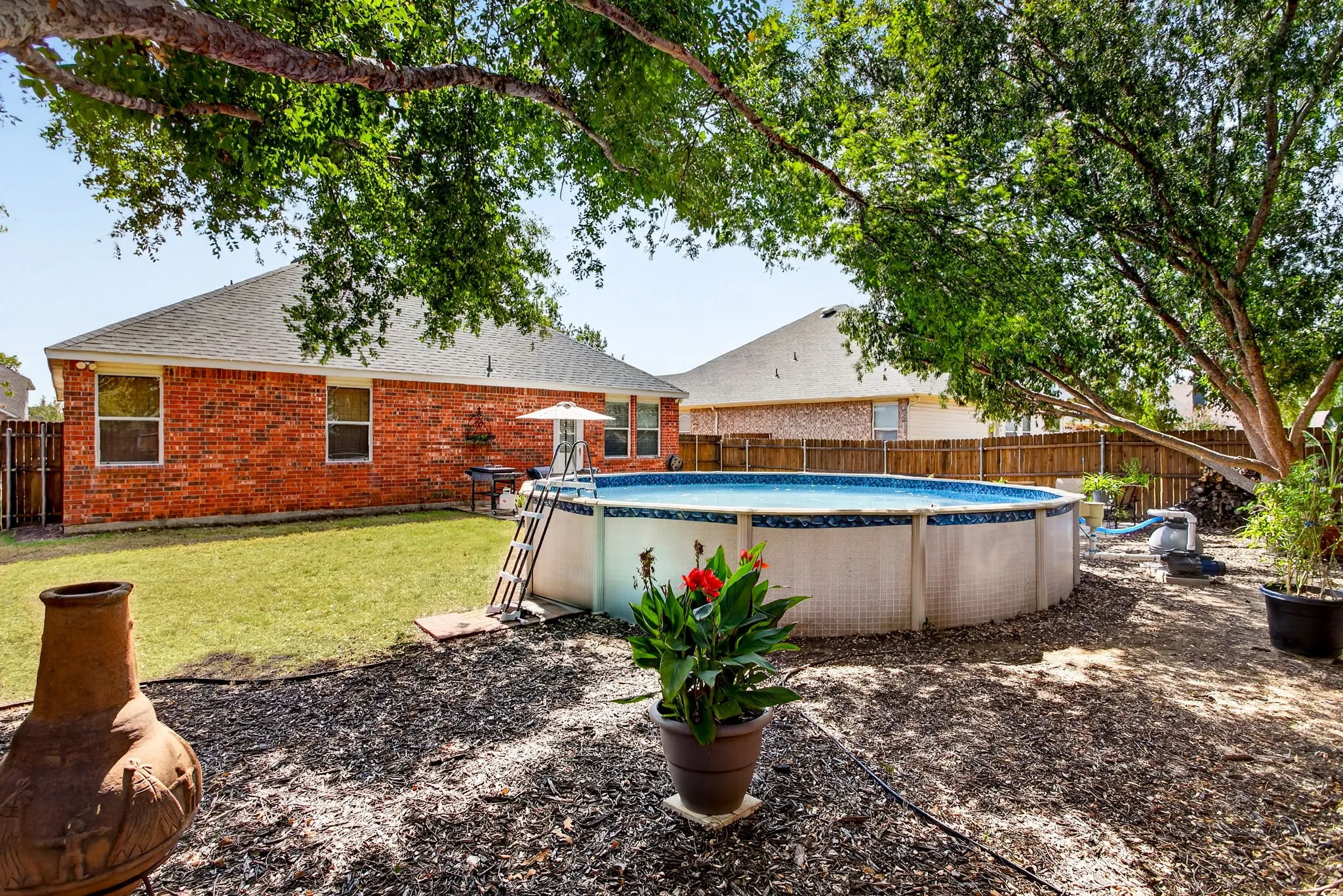 Expansive backyard featuring a brick exterior, an above-ground swimming pool, a mature shade tree, and a partially manicured lawn