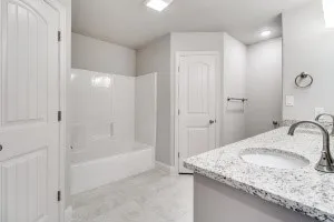 White alcove bathtub with integrated shower, dual white paneled doors, light grey wall paint, grey tile flooring, and a vanity featuring a light-toned countertop with integrated sink