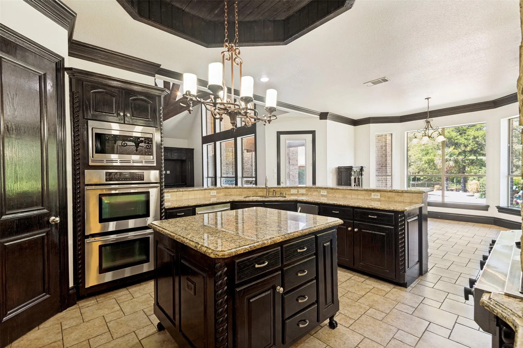Kitchen featuring dark wood cabinetry, light-toned stone countertops, stainless steel double ovens, tile flooring, and a decorative chandelier