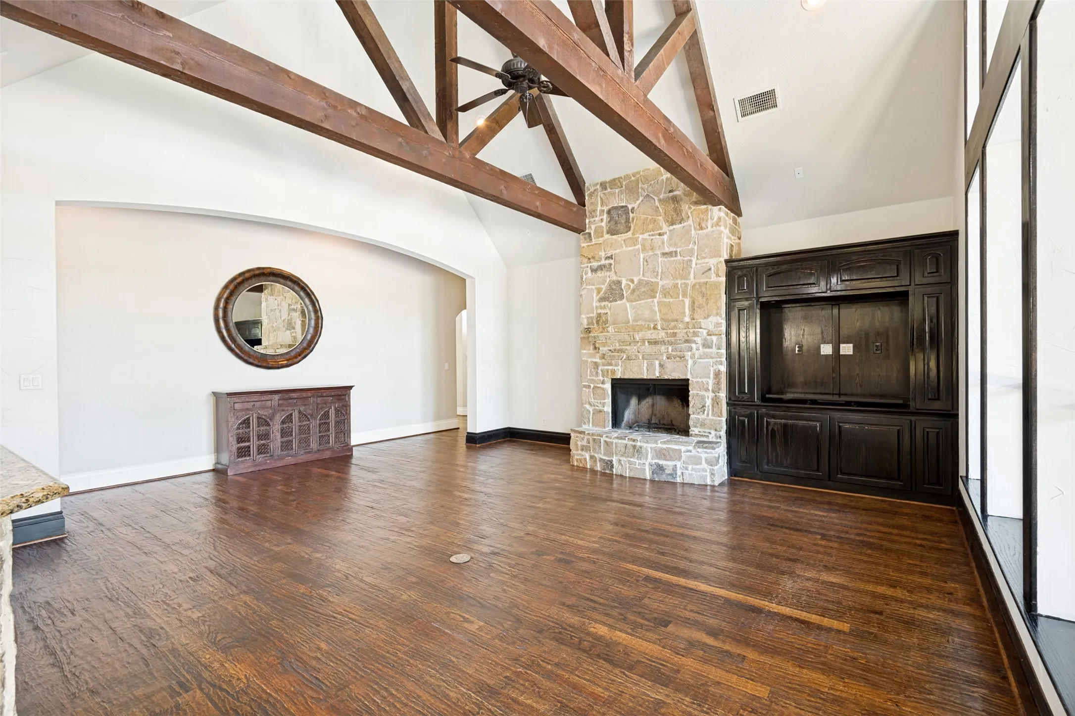 Vaulted ceiling with exposed wood beams, stone fireplace, built-in dark wood cabinetry, wood-finish flooring, and an arched wall opening