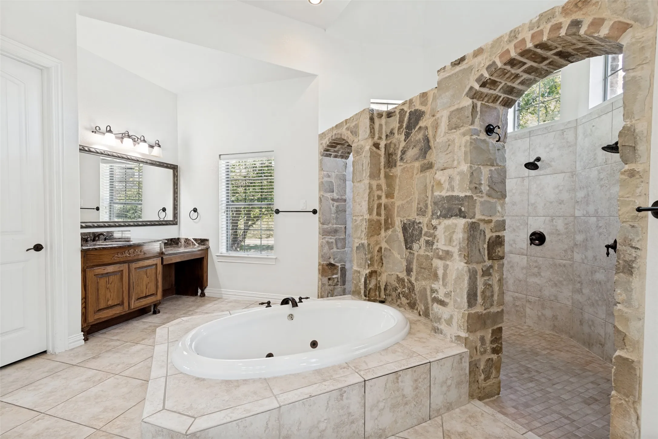 Arched stone wall, walk-in shower with multiple showerheads, built-in soaking tub, wood-finish vanity with dual sinks, and tile flooring