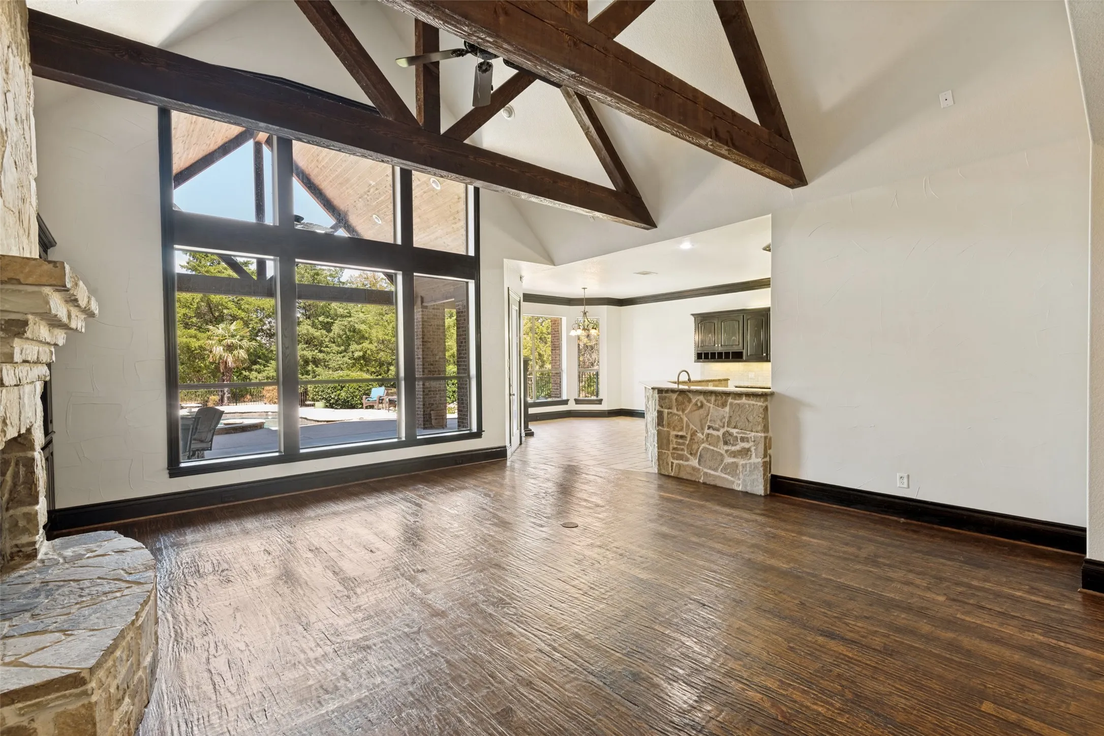 Vaulted ceiling with exposed wood beams, stone-clad fireplace, full-height windows, wood-finish flooring, and built-in wet bar with stone surround