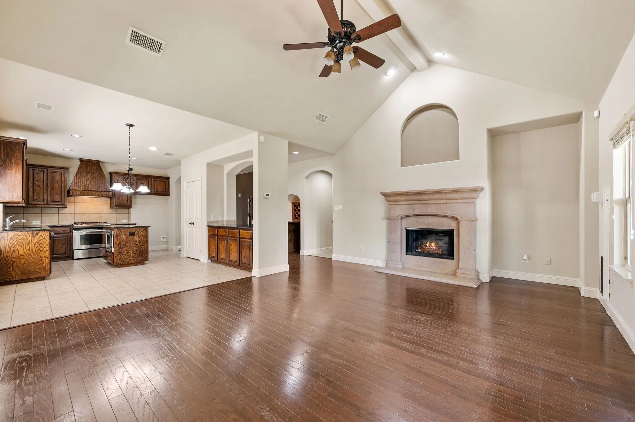 Vaulted ceiling with wood-finish fan, stone-surround fireplace, wood-finish flooring, and kitchen with wood​​‌​​​​‌​​‌‌​‌‌​​​‌‌​‌​‌​‌​​​‌​​ cabinetry