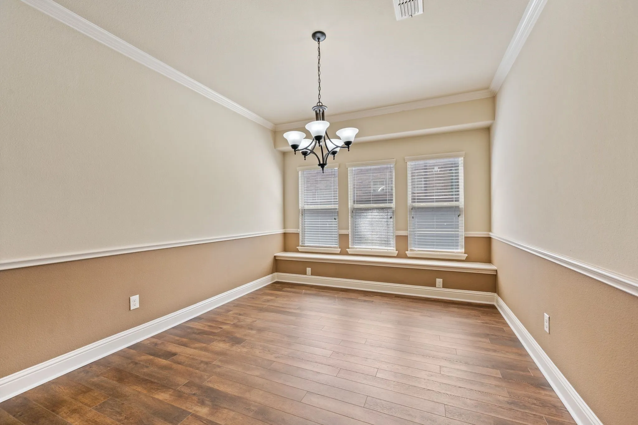 Wood-finish flooring, three windows with blinds, a chandelier with white glass shades, crown molding, and two-tone wall treatment