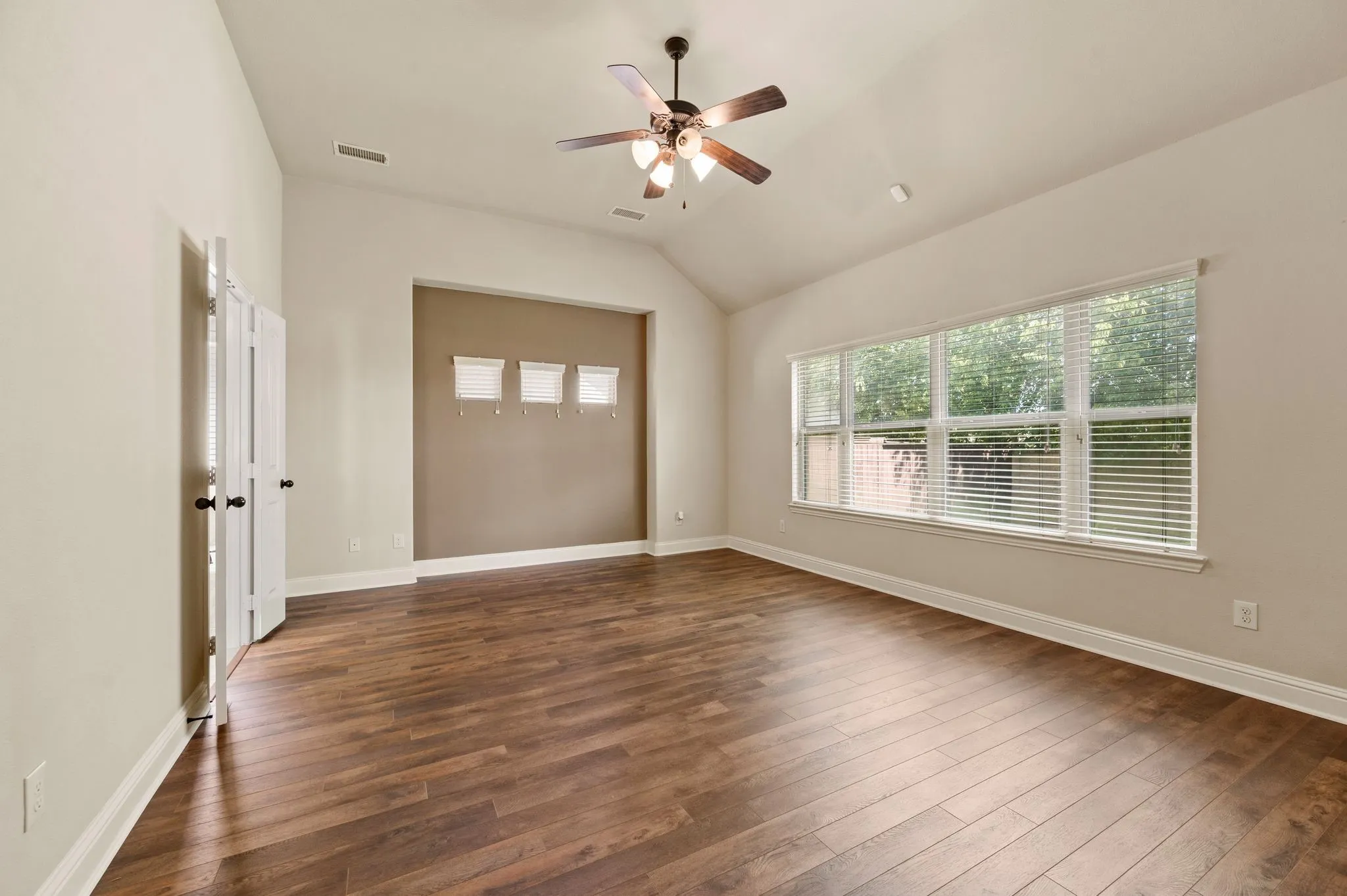Wood-finish flooring, vaulted ceiling, expansive window with blinds, ceiling fan with light, and white paneled bi-fold doors