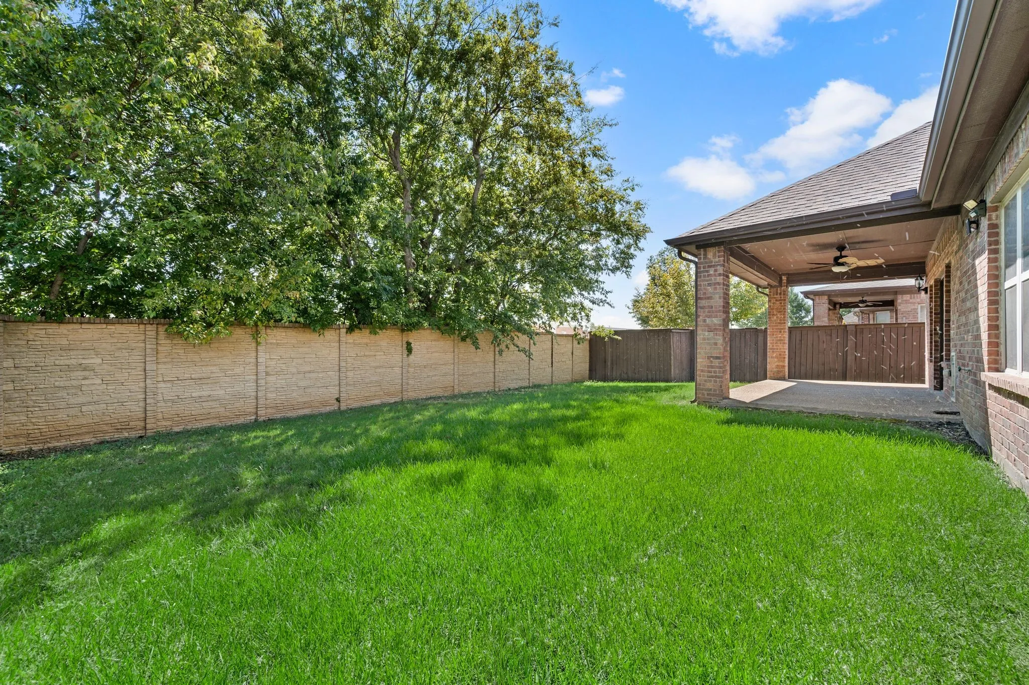Expansive backyard featuring a manicured lawn, a full-height stone-finish fence, and a covered patio with brick columns