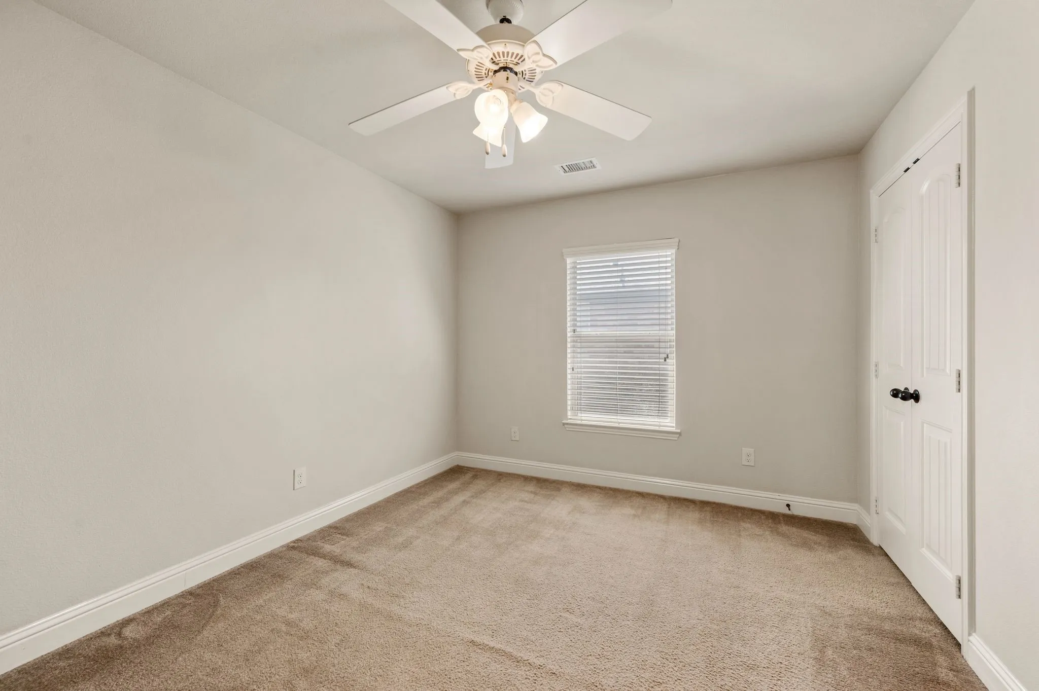 Neutral wall paint, carpeted flooring, window with blinds, ceiling fan with integrated lighting, and a paneled door with dark hardware