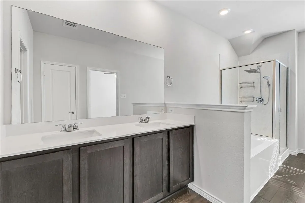 Dual vanity with integrated sinks, expansive mirror, dark wood-finish cabinetry, separate glass-enclosed shower, and built-in bathtub