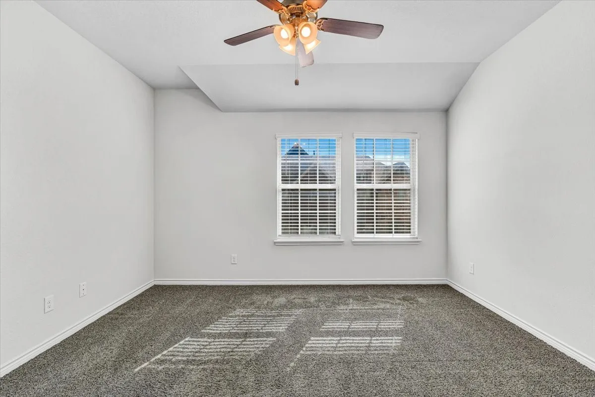 Recessed tray ceiling, neutral wall paint, ceiling fan with integrated lighting, dual windows with blinds, and gray carpeting