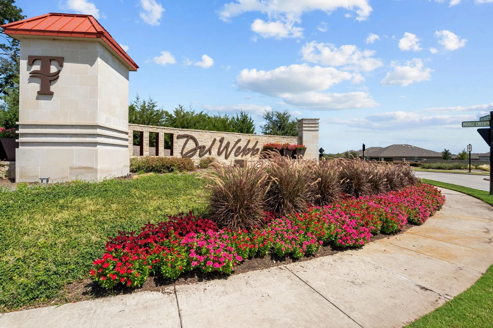 Community entrance featuring a stone monument with a red tile roof, decorative landscaping with flowering plants and ornamental grasses, and a paved sidewalk