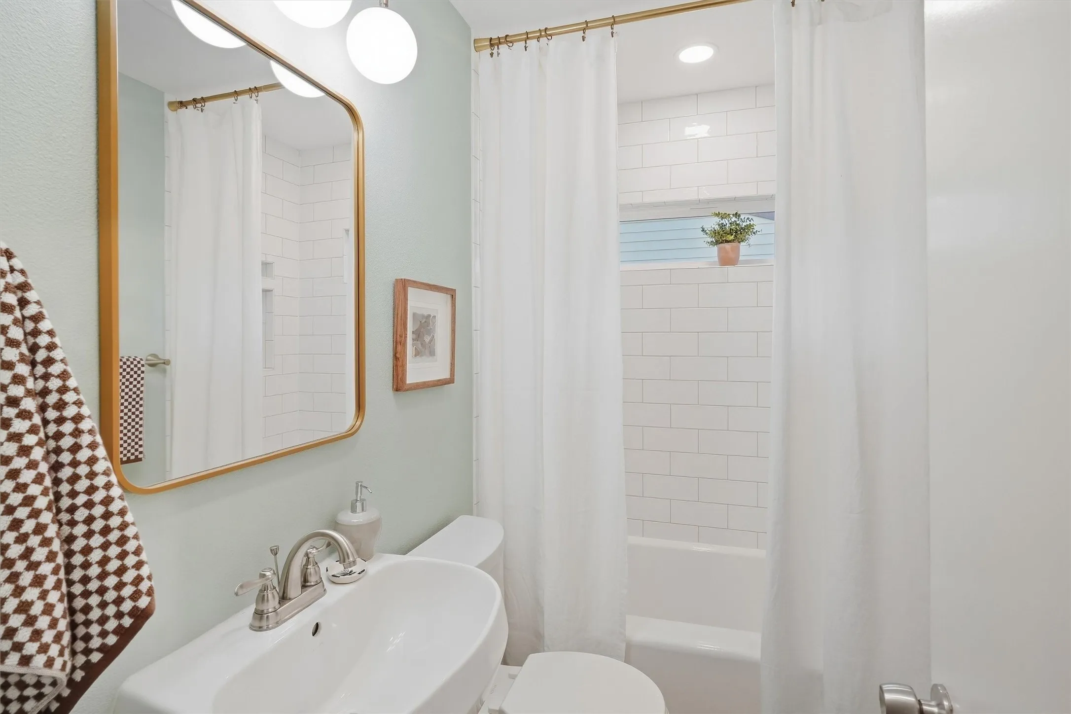 The updated full secondary bathroom highlights soft sage-green walls, a curved white pedestal sink, and stylish warm-gold accents including a arched mirror frame and curtain hardware. A tiled tub-and-shower combination features crisp white subway tile and a high accent window for privacy and natural light.