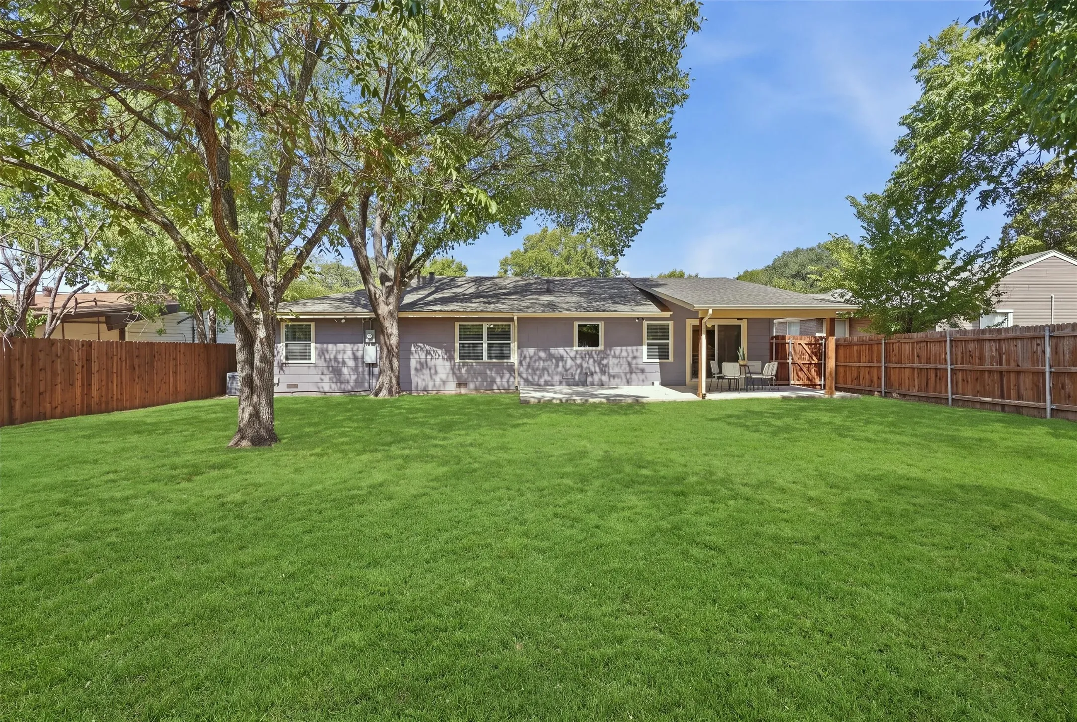 The expansive, fully fenced backyard boasts a vibrant green lawn shaded by mature trees and a comprehensive wood privacy fence. A covered back patio connects directly to the home, creating an ideal outdoor setting for entertaining, pets, or play.