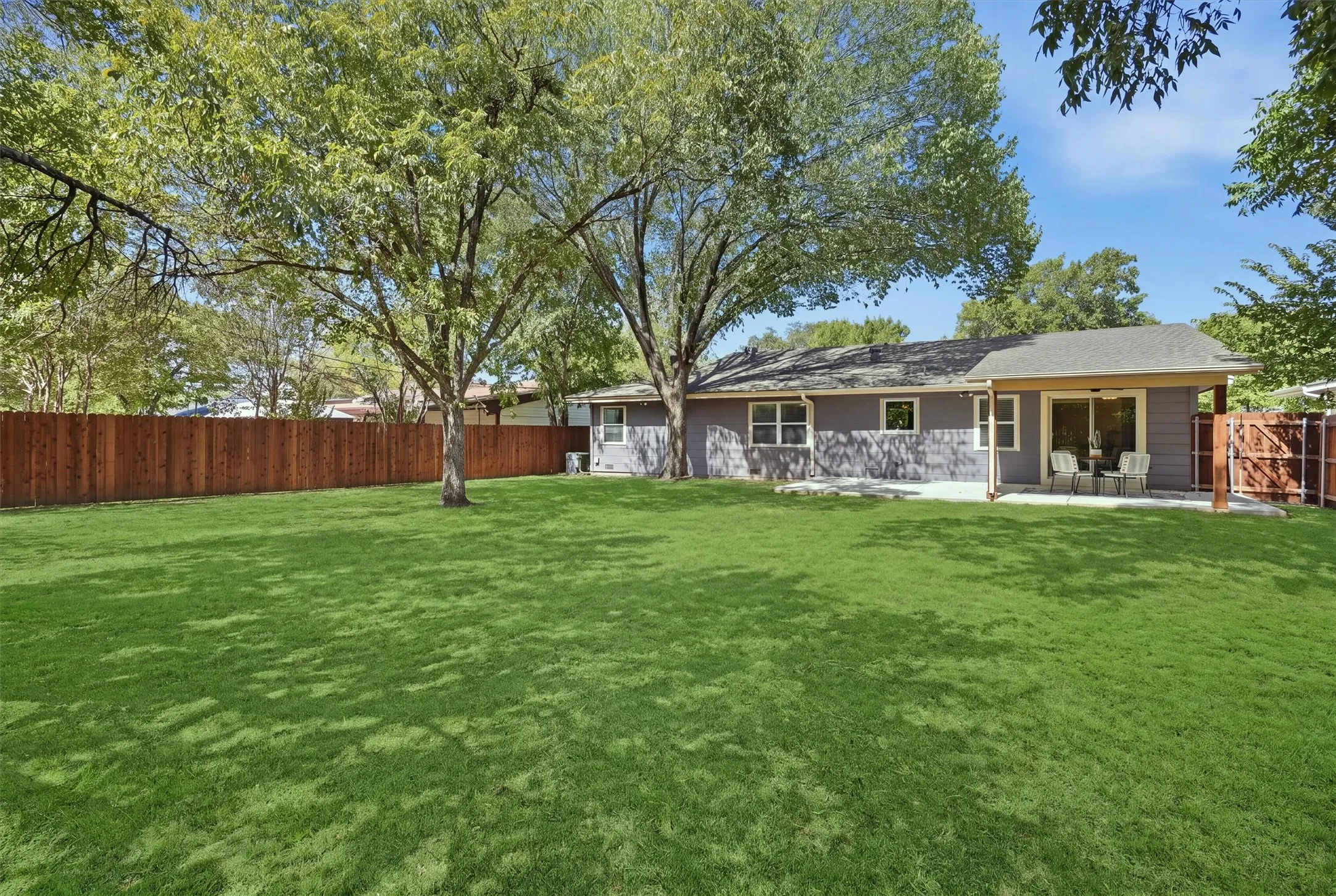 An alternate view of the backyard highlights the impressive depth of the lawn, surrounded by sturdy wooden privacy fencing on all sides. Beautiful mature trees cast dappled shade across the property, providing a serene outdoor escape with plenty of space for recreation.