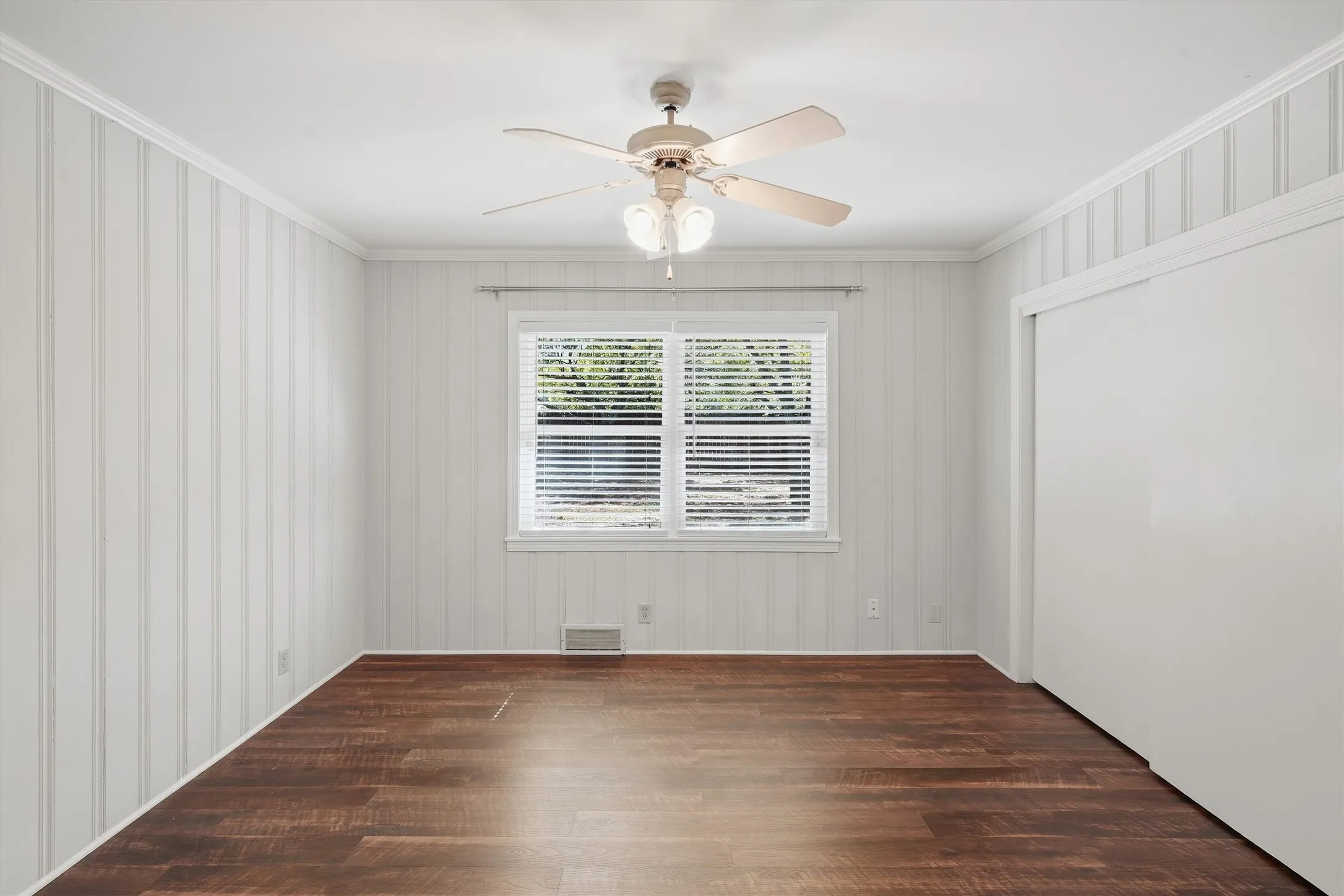 This additional secondary bedroom features vertical beadboard wall detailing, rich wood-look flooring, and a central ceiling fan with light fixture. A wide window with blinds provides bright natural light, while a large sliding-door closet offers ample storage space.