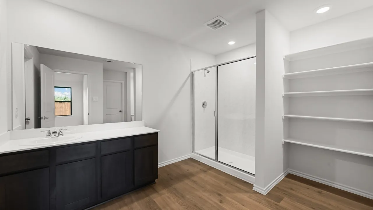 Bathroom featuring a single-sink vanity with dark cabinetry, an elongated mirror, a clear glass shower enclosure, wood-finish flooring, and built-in shelving